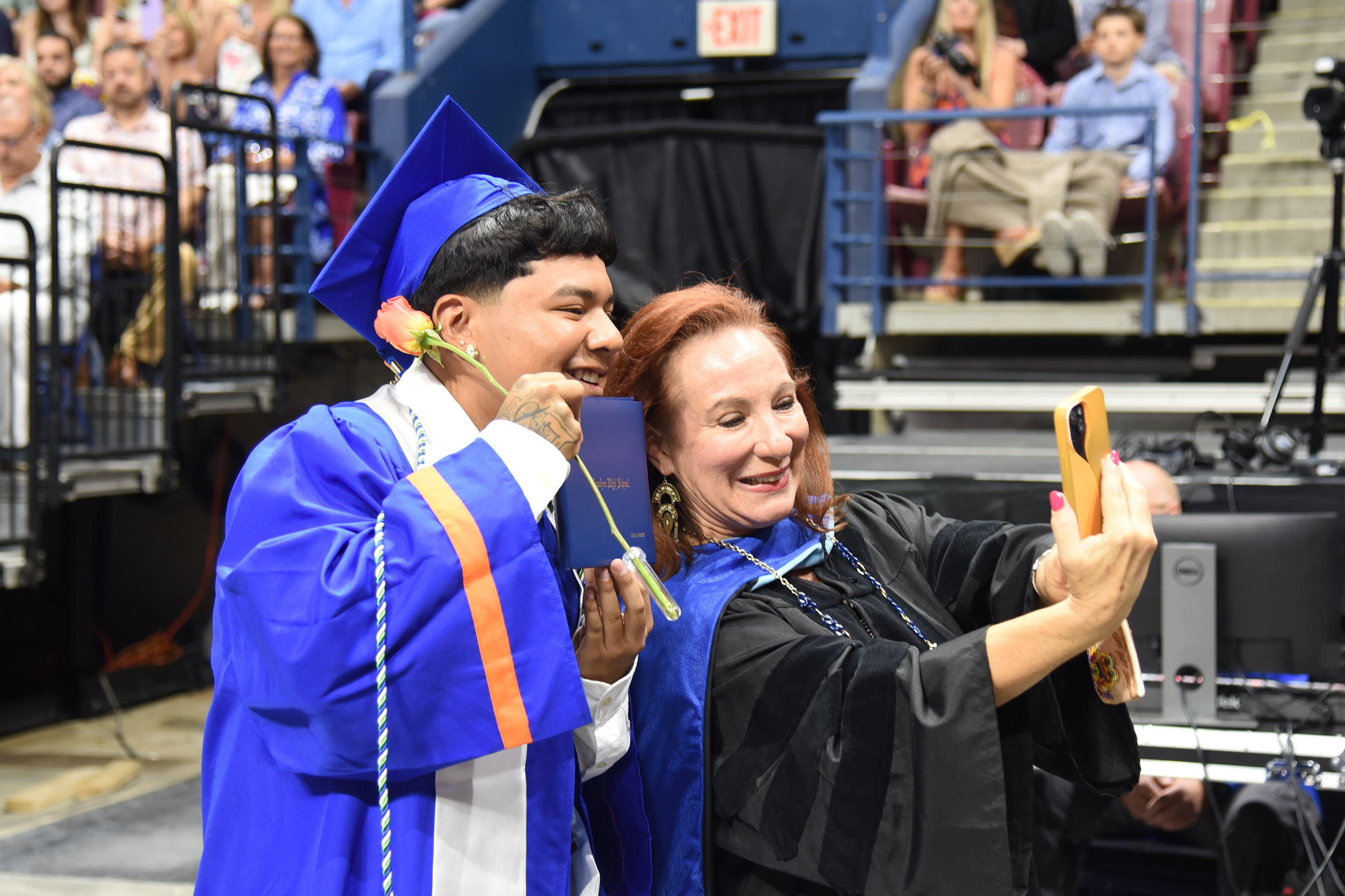 A young man in a blue graduation robe kisses a woman wearing a black robe during a ceremony.