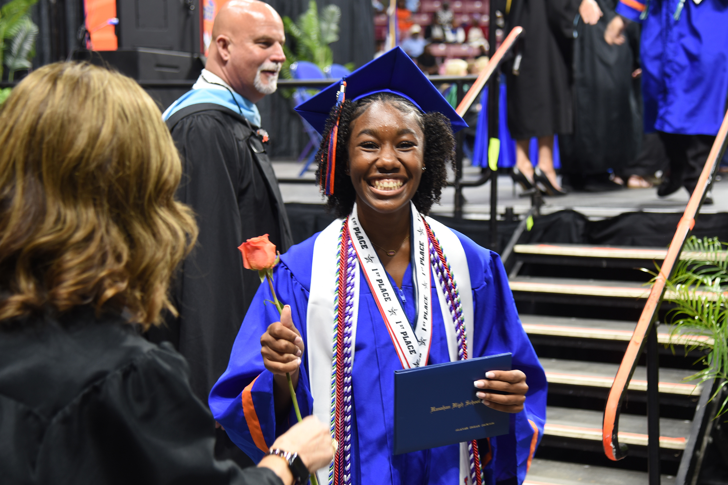 A woman in a blue gown and cap smiles at a person holding a rose. Behind them, a stage with people in blue gowns.