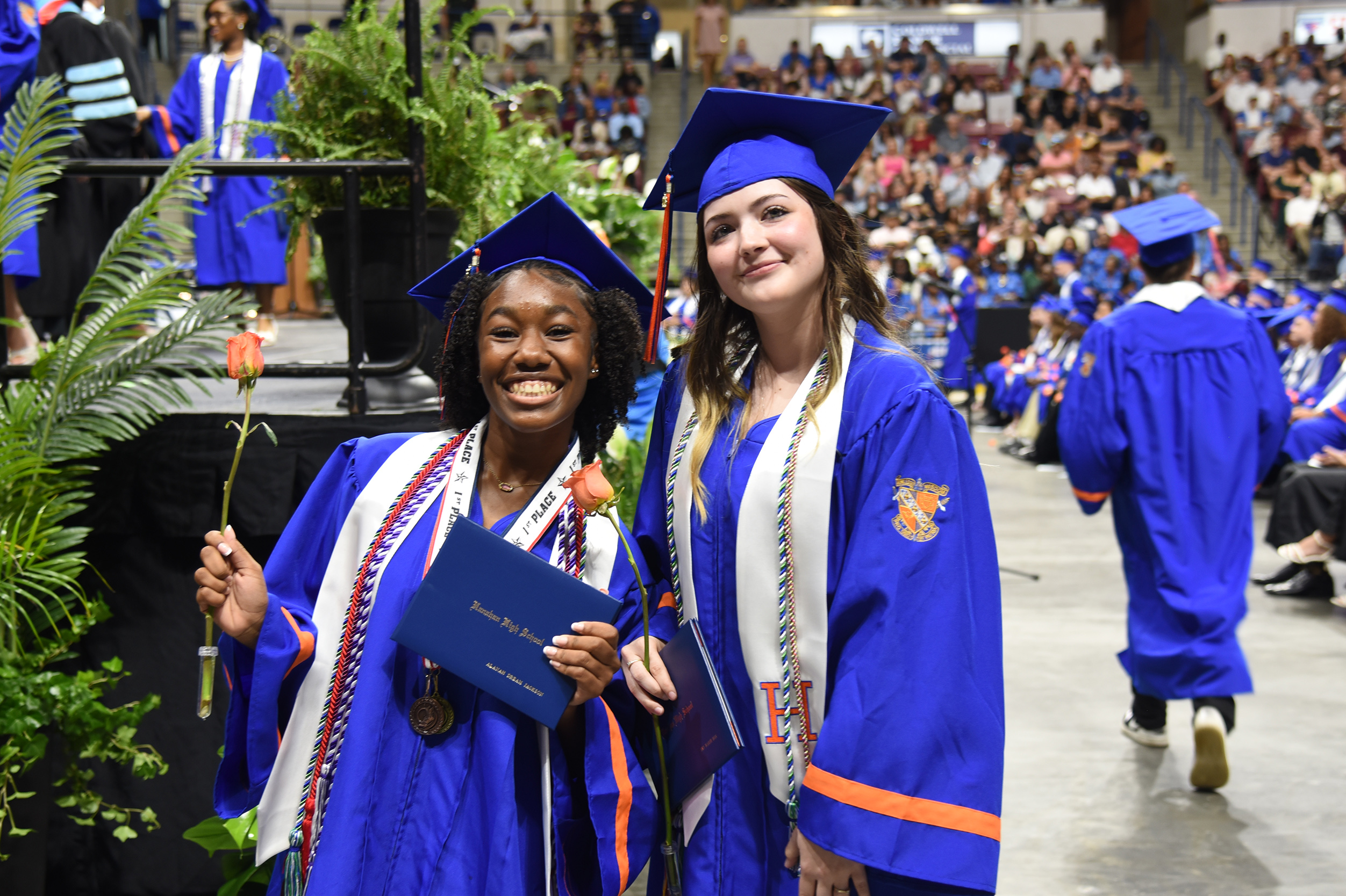 Two women in blue caps and gowns, one holding a blue folder and a rose, smiling at a school event.