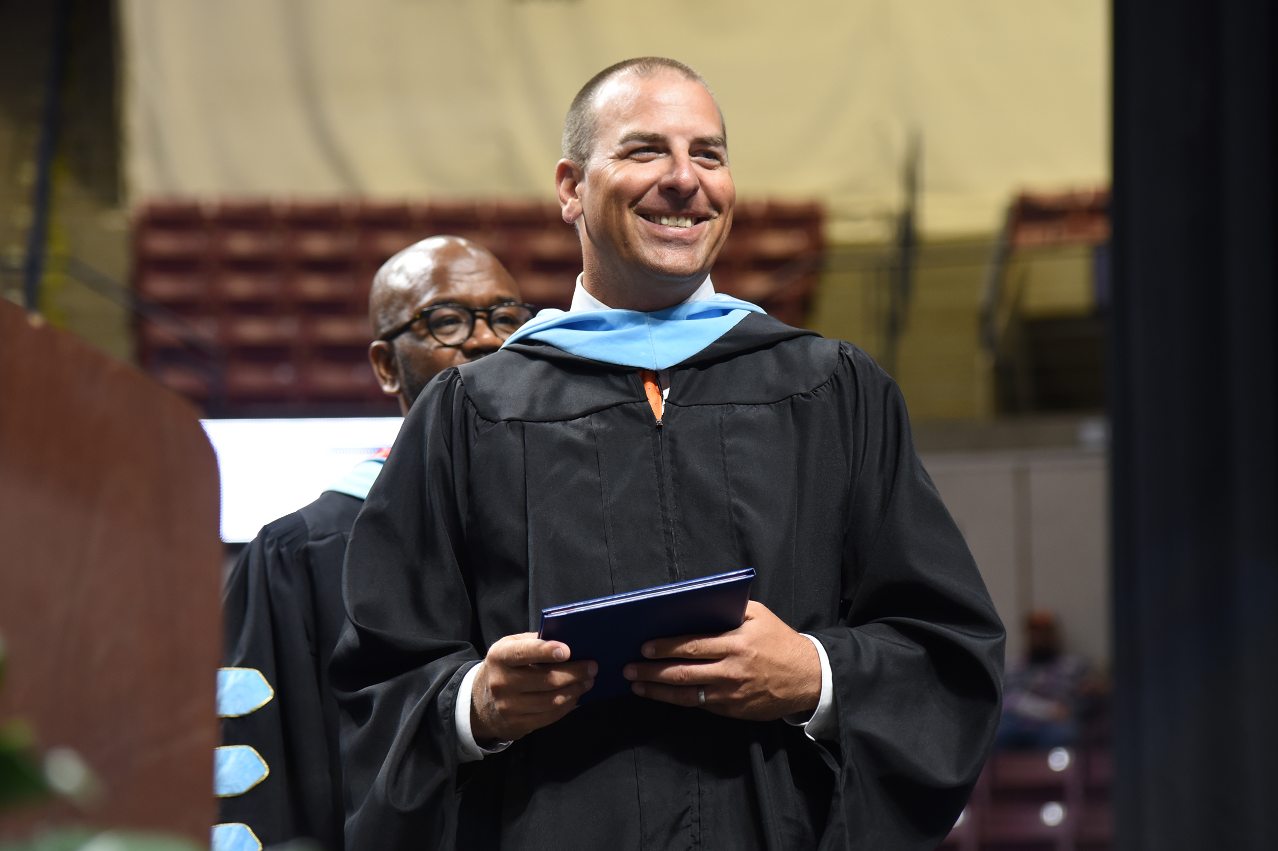 A person in a graduation gown holding a diploma and smiling in an indoor auditorium with empty chairs in the background.