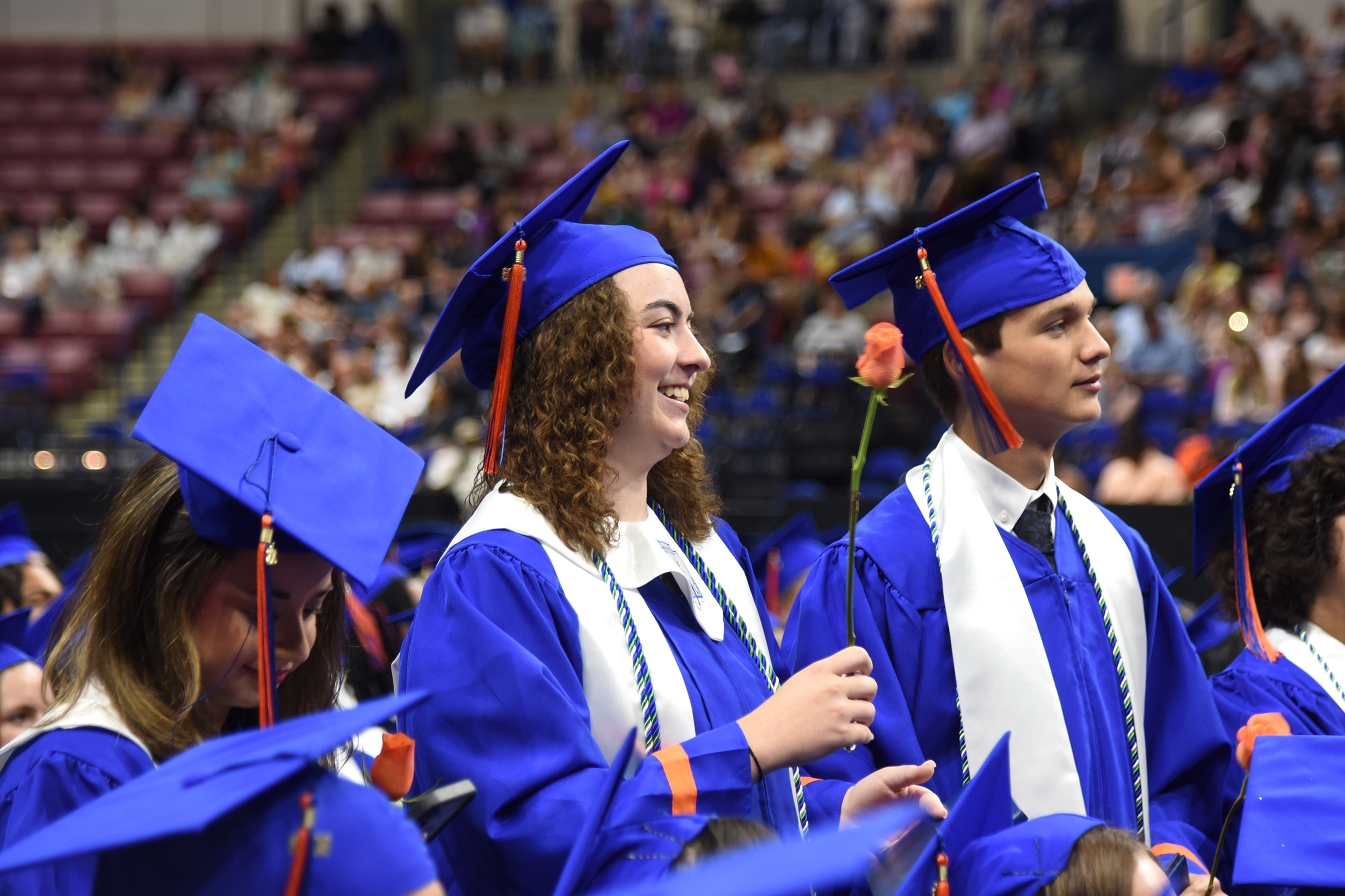 Graduates in caps and gowns, some holding flowers, standing in an auditorium. People seated in rows in the background.