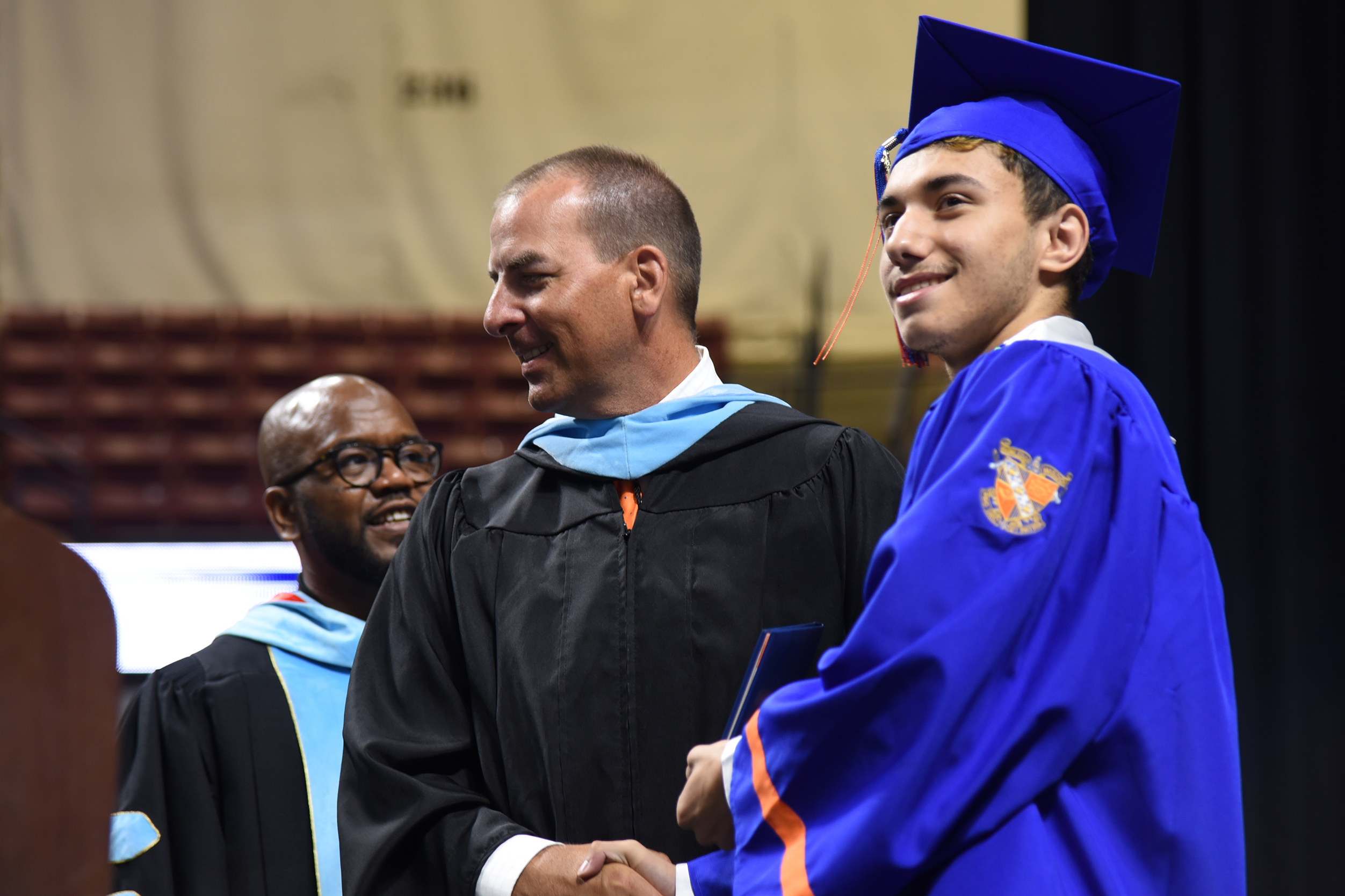 A man shakes hands with a graduate at a ceremony. The graduate wears a blue robe. Behind them, another man wears glasses.