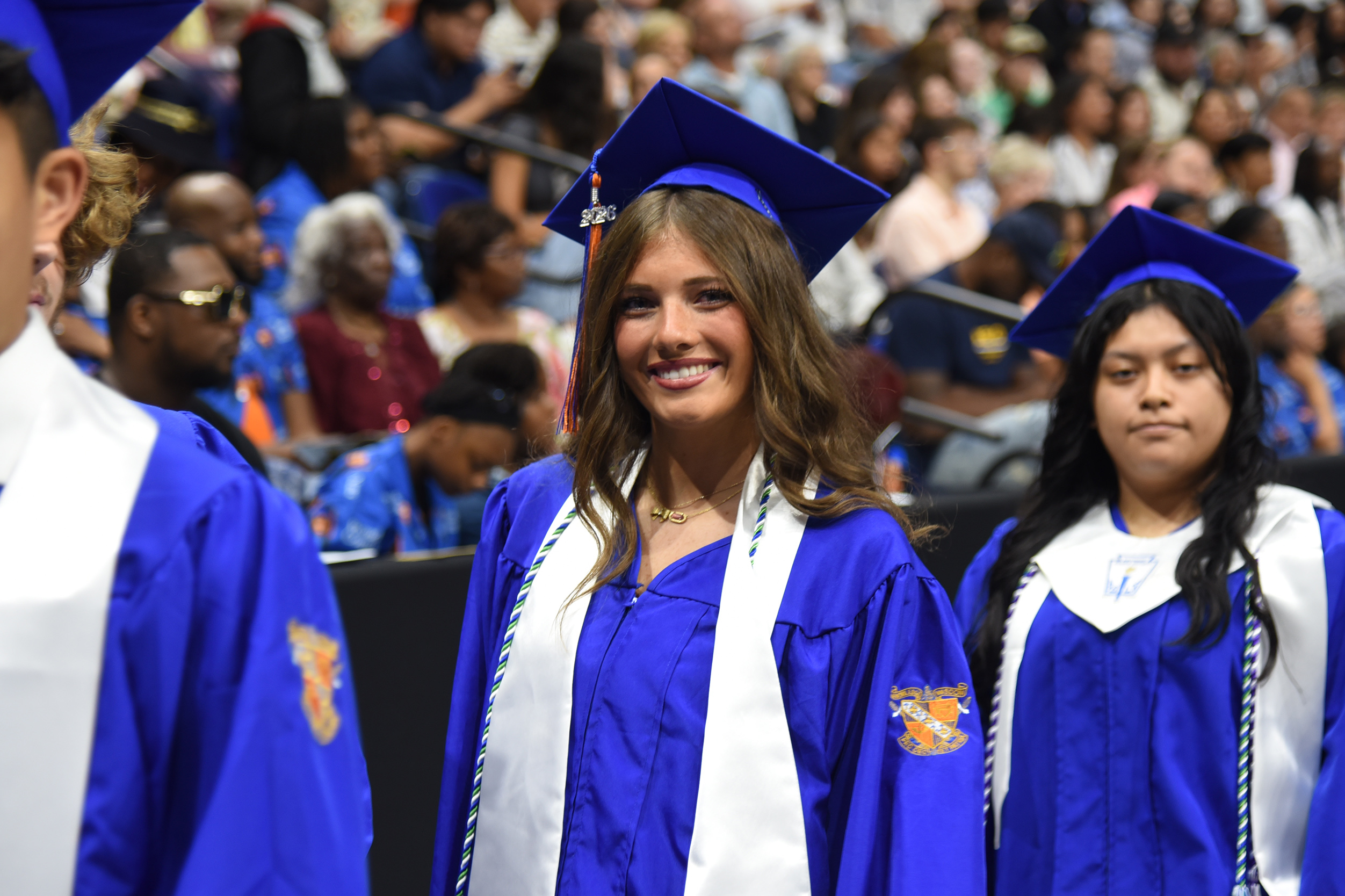 Three graduates in blue gowns and caps stand in front of a large crowd, smiling.