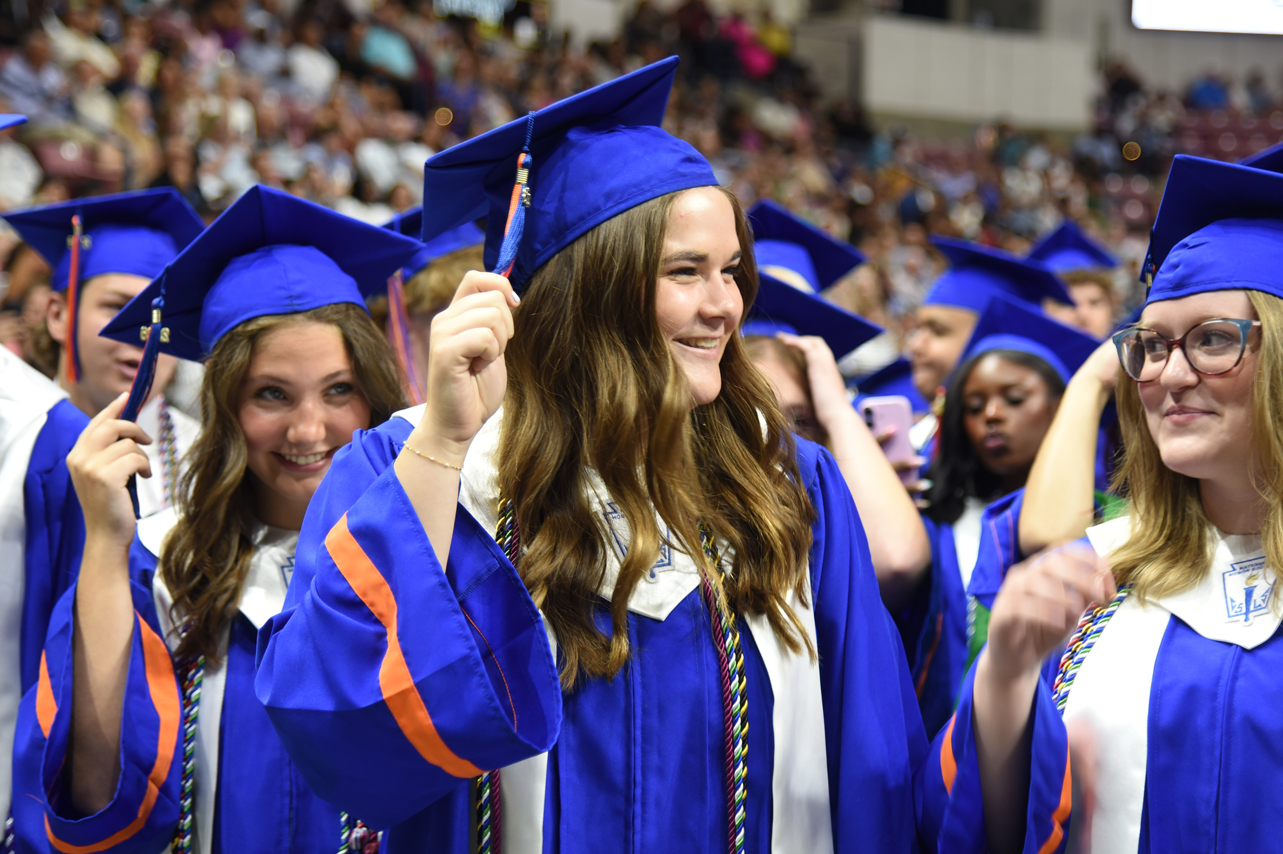 A group of graduates in blue gowns with orange and white sashes at a ceremony.