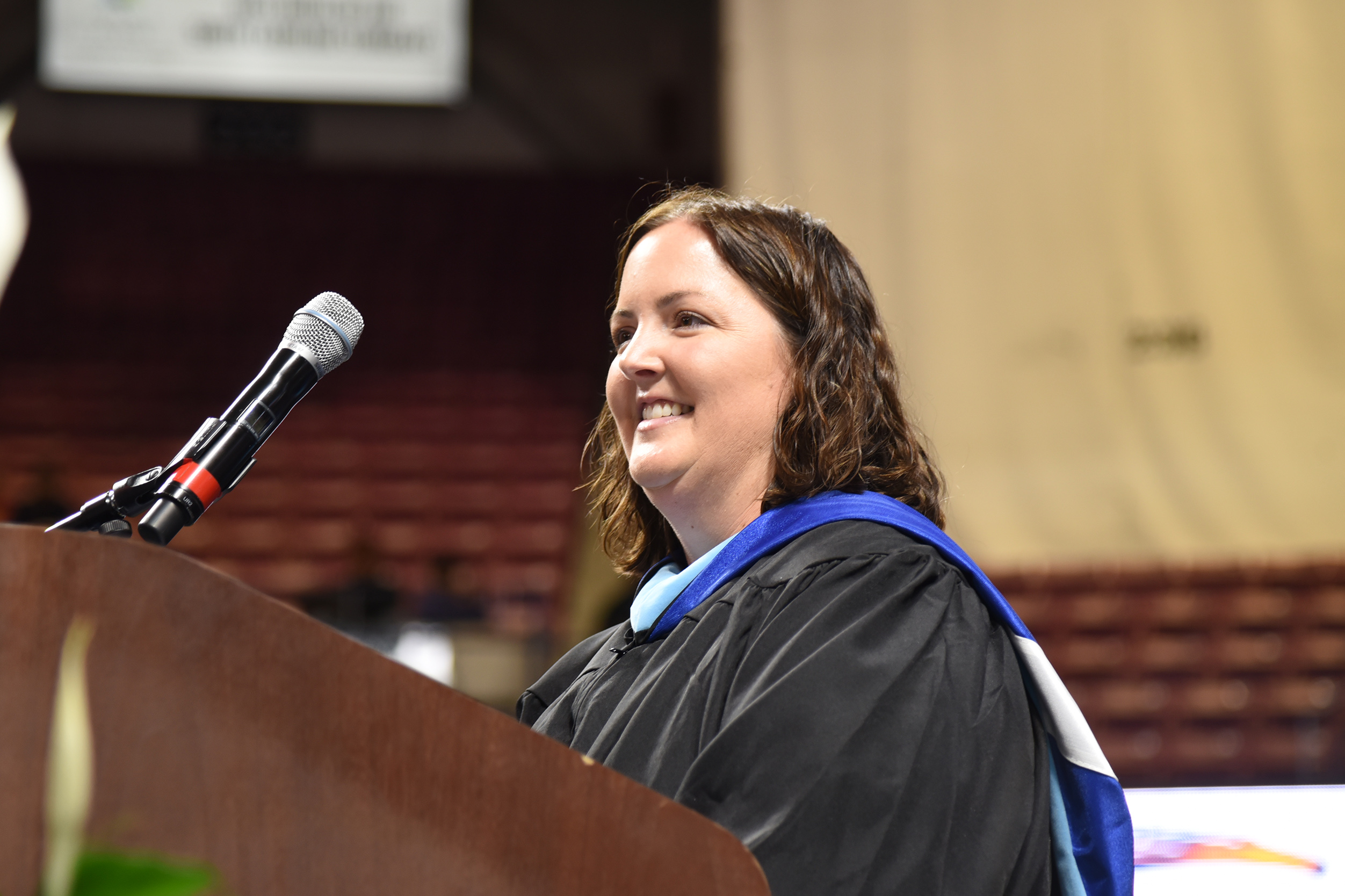 A woman with a smiling face, wearing a graduation gown, stands at a podium with a microphone.