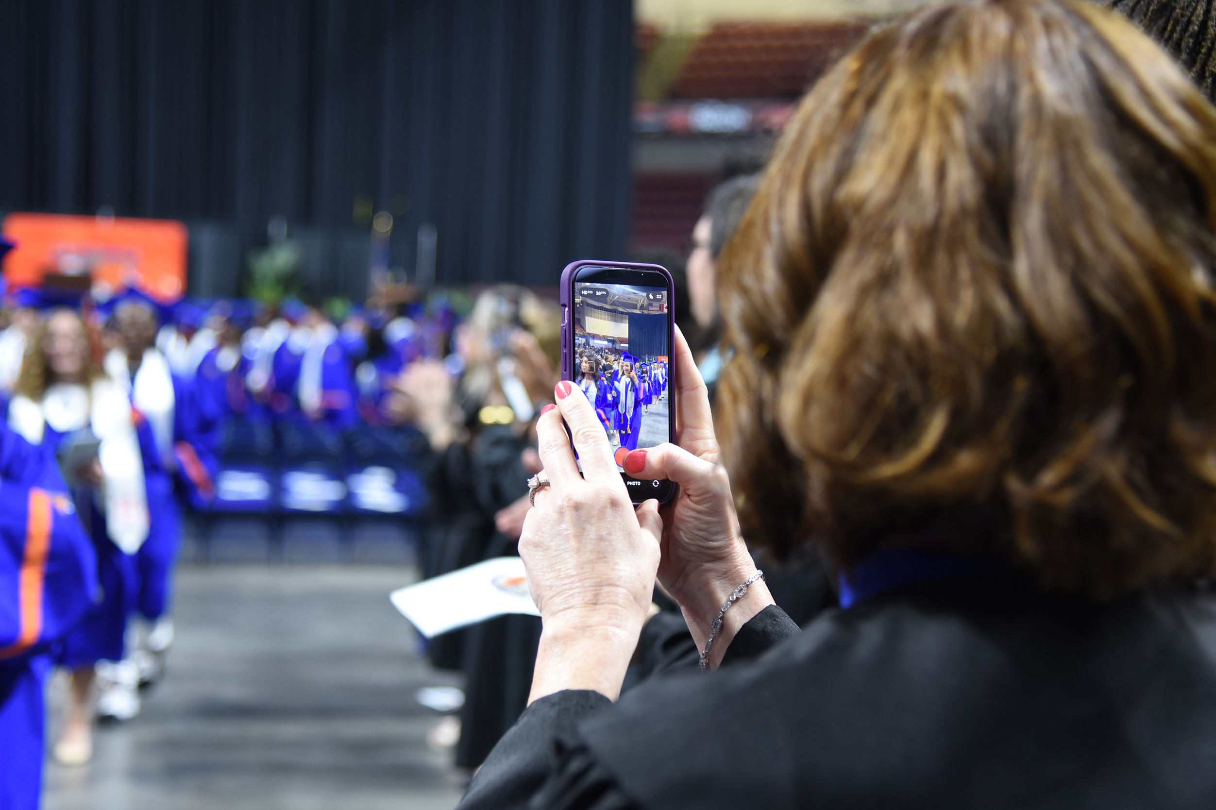 A woman with brown hair in a black outfit holds a phone and takes a photo of a graduation ceremony.