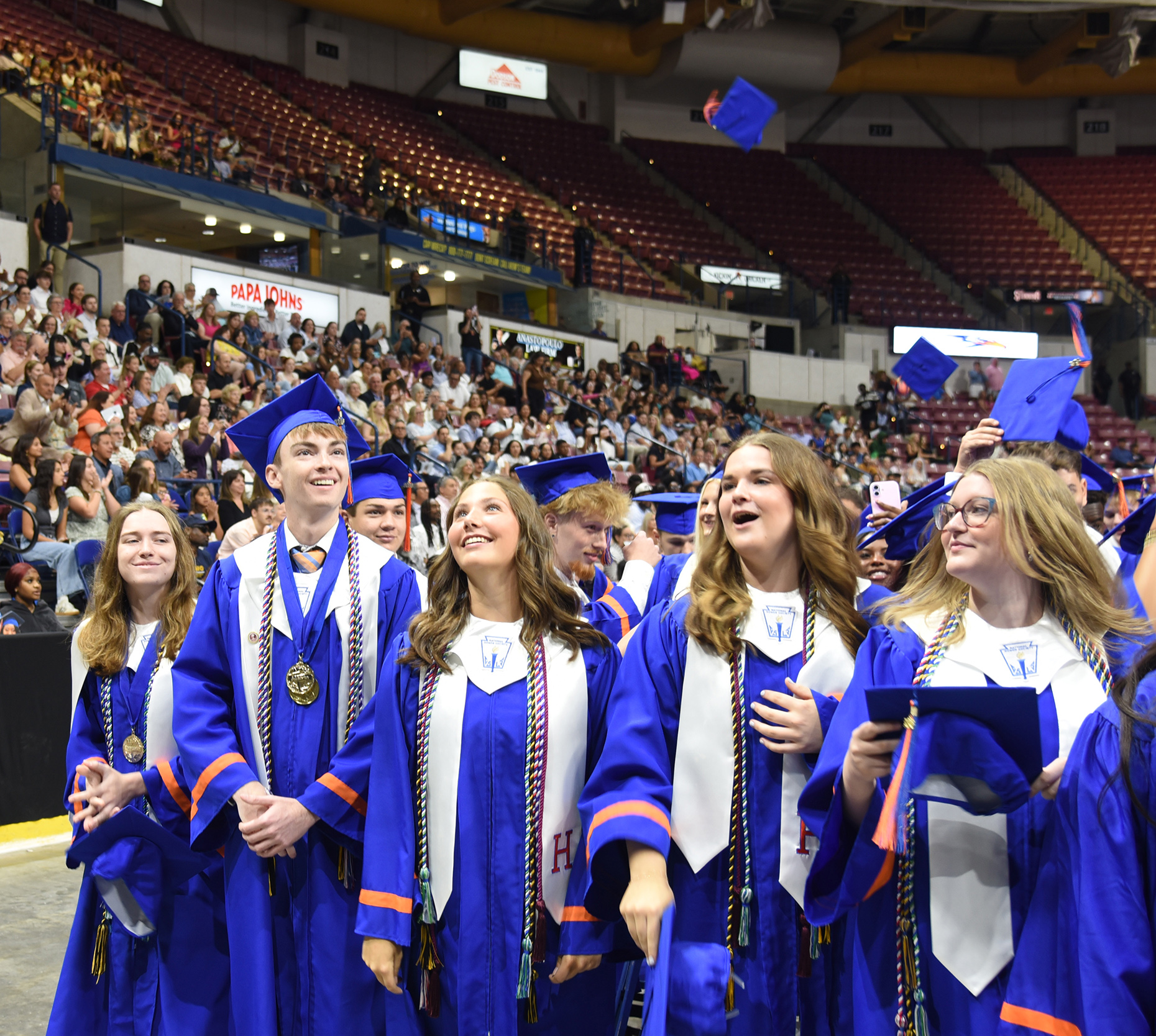 Graduates in blue gowns and caps stand together in an arena with a cheering crowd in the background.