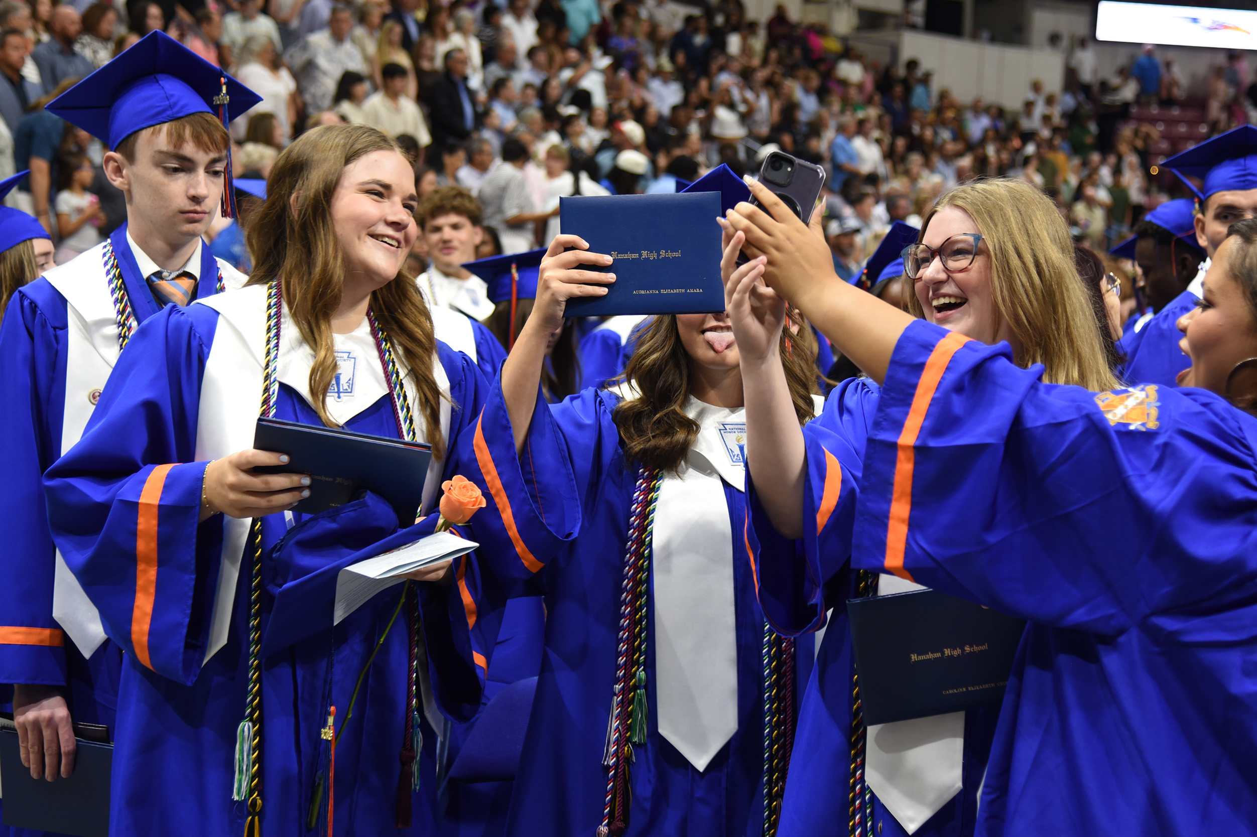 Graduates in blue gowns and caps celebrate, one holding a diploma, while another lifts a certificate in a large, crowded room.