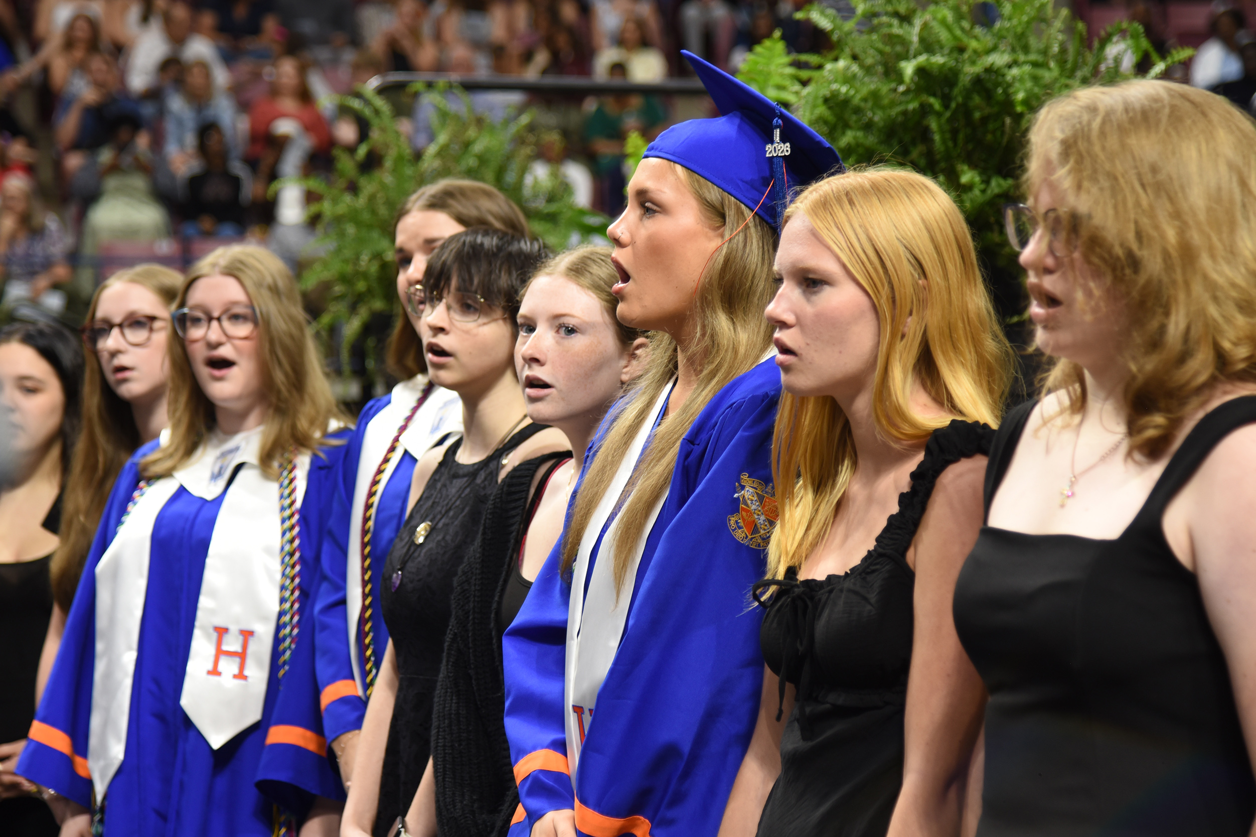 A group of women in blue and white robes and black dresses, possibly singing, in an outdoor setting.