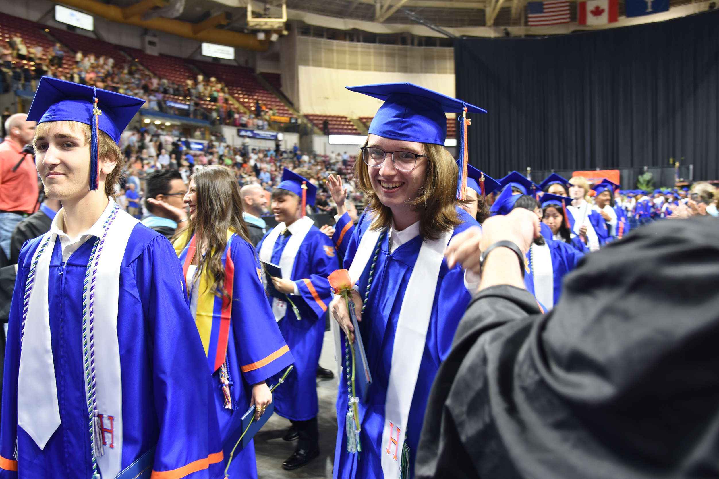 Graduates in blue gowns and caps walk down the aisle at a ceremony. An adult shakes hands with a graduate.