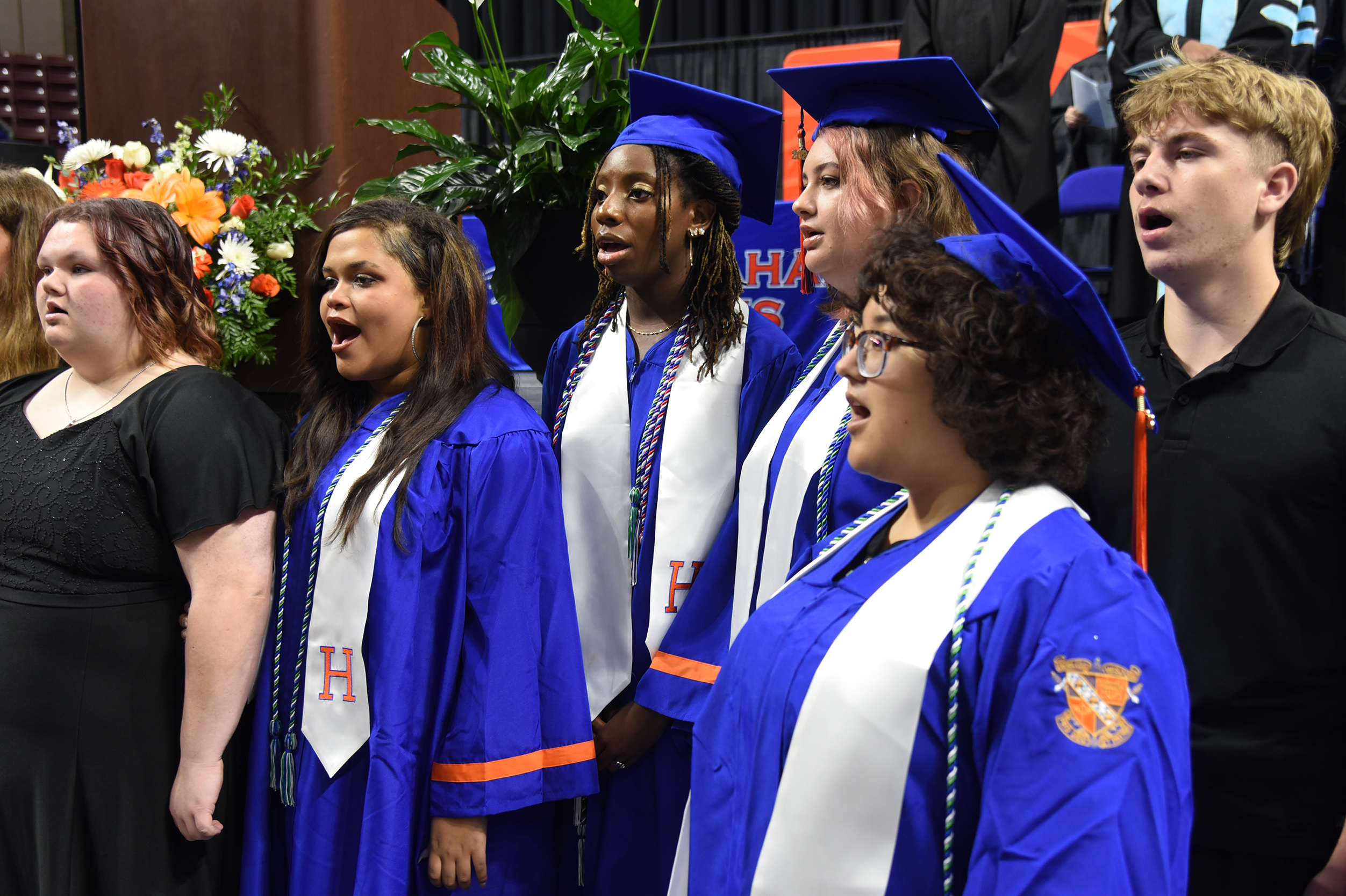 Group of students in blue and white graduation gowns sing, accompanied by a woman in a black dress.