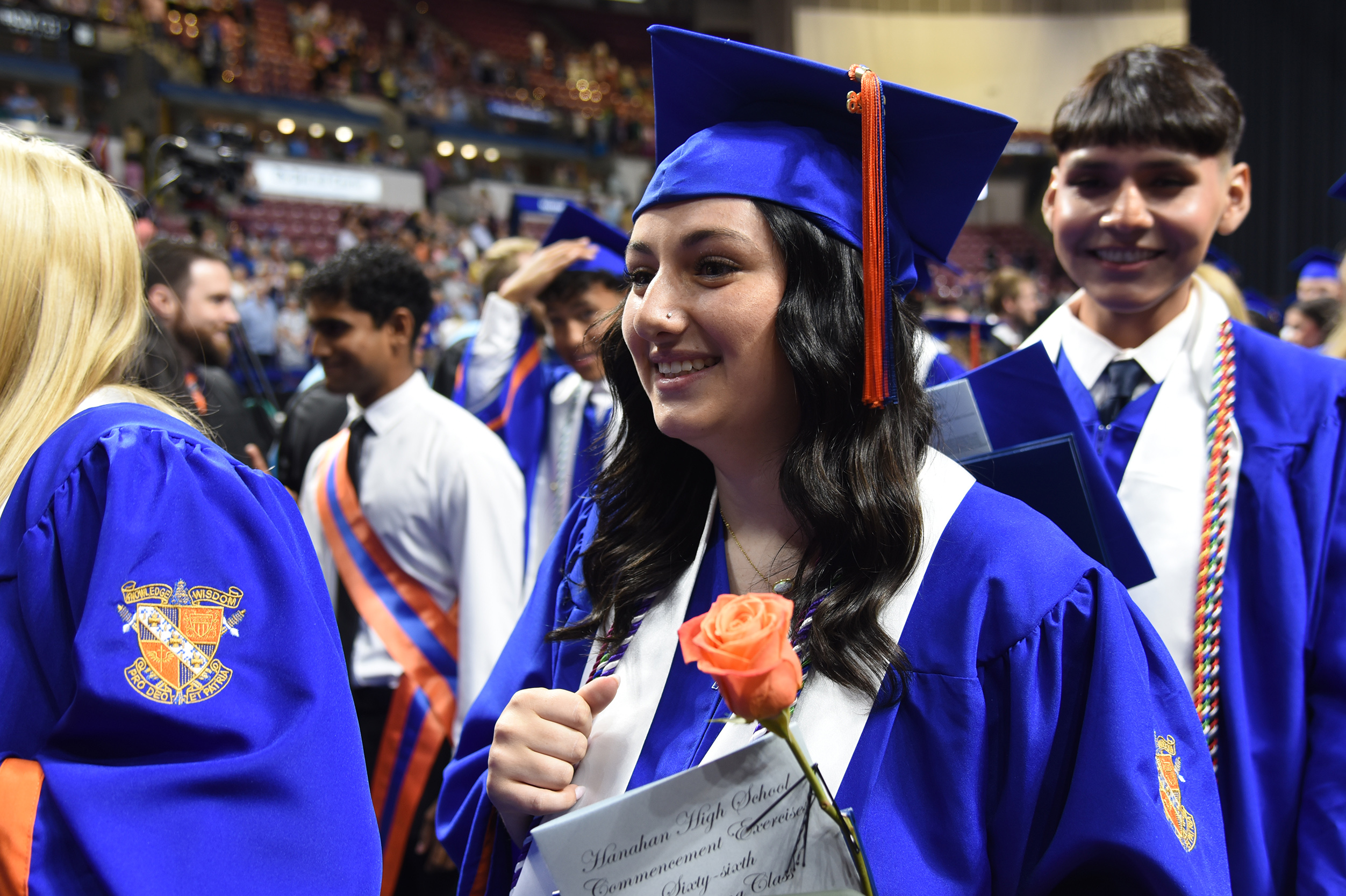 Graduates in blue and white gowns and caps, one smiling with a rose, at an indoor event.