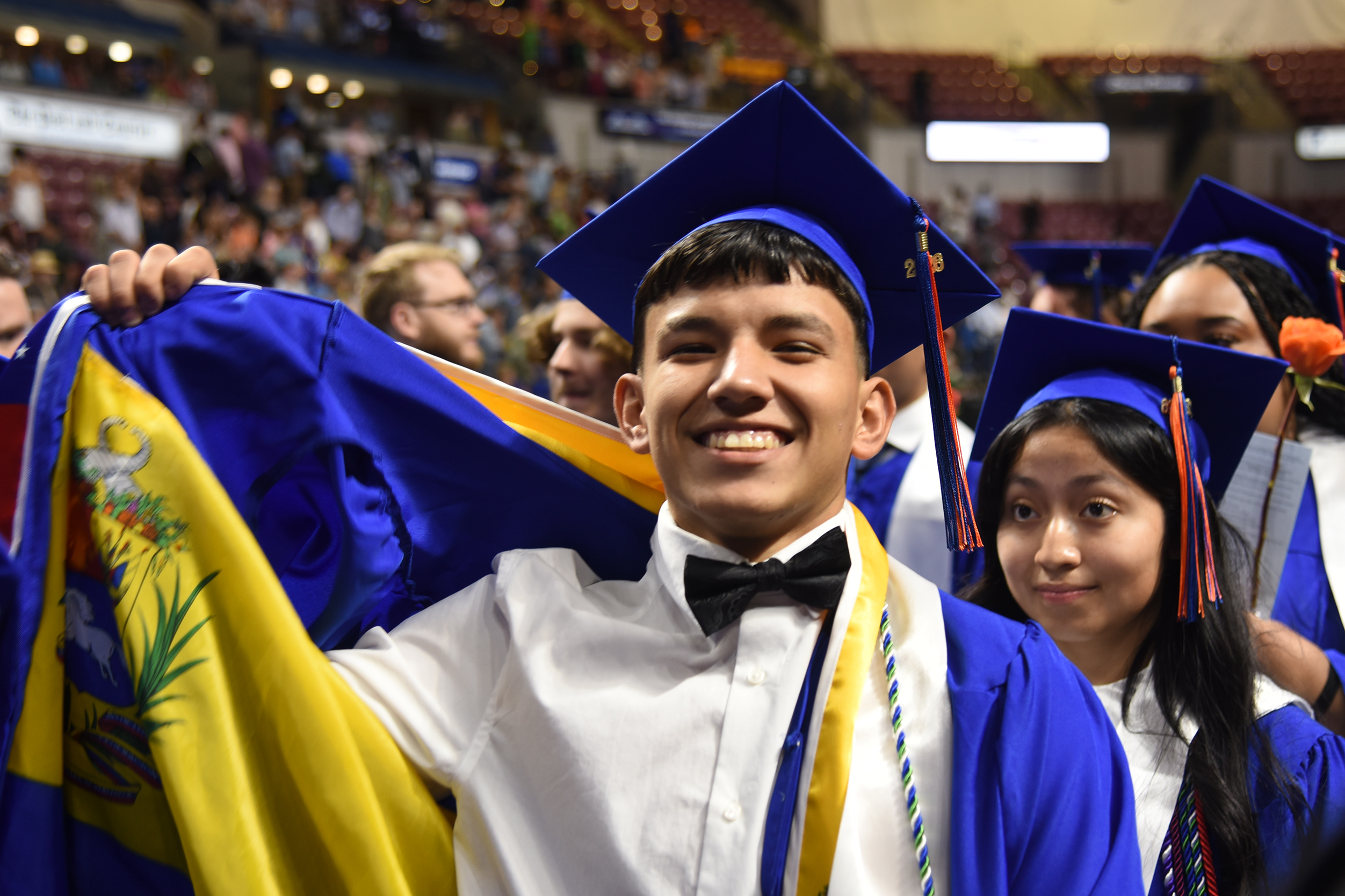 A smiling man in a graduation gown holds a flag while standing in front of a crowd.