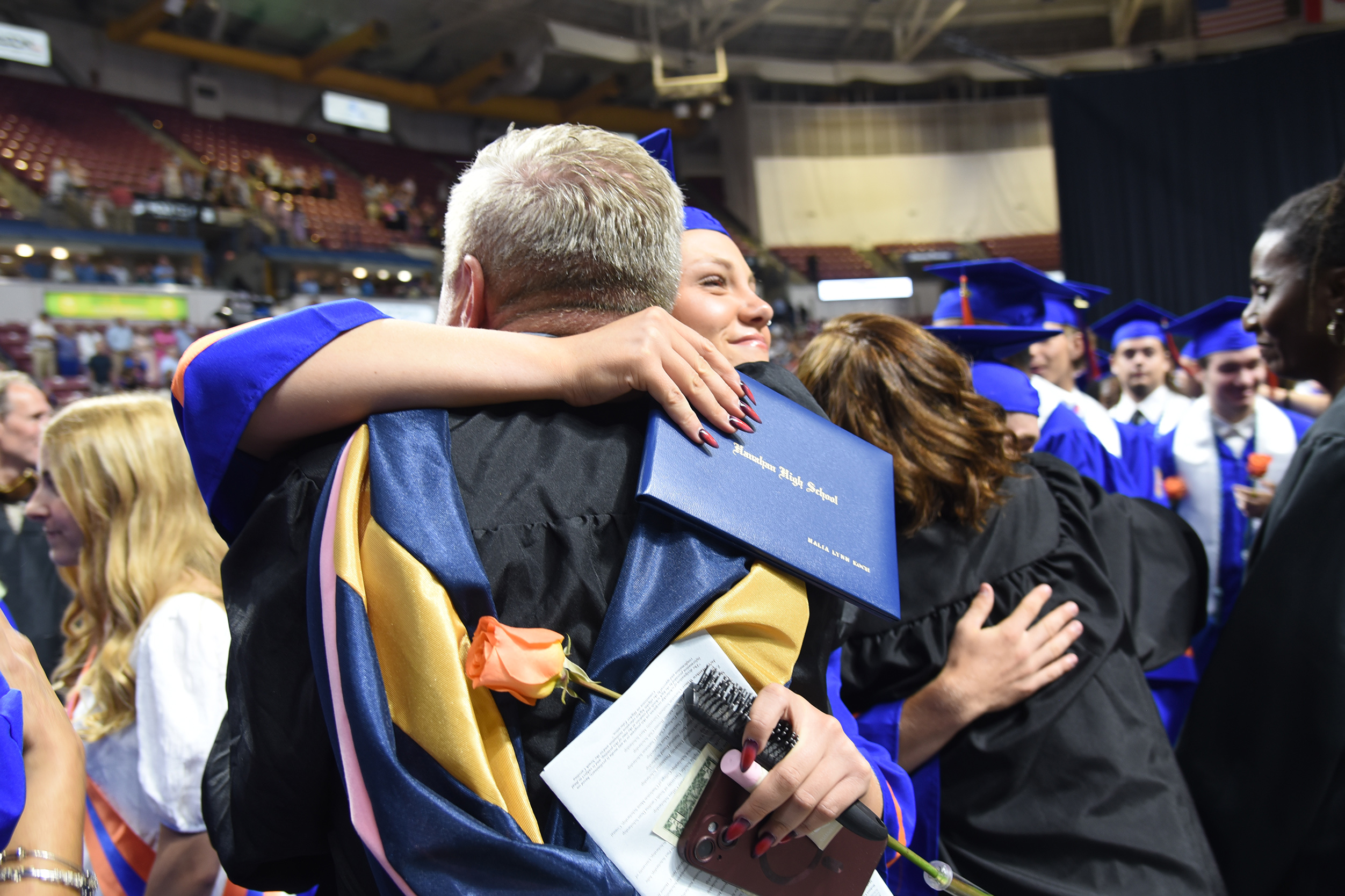A man in a cap and gown embraces a graduate while holding a diploma, surrounded by others in a crowded auditorium.