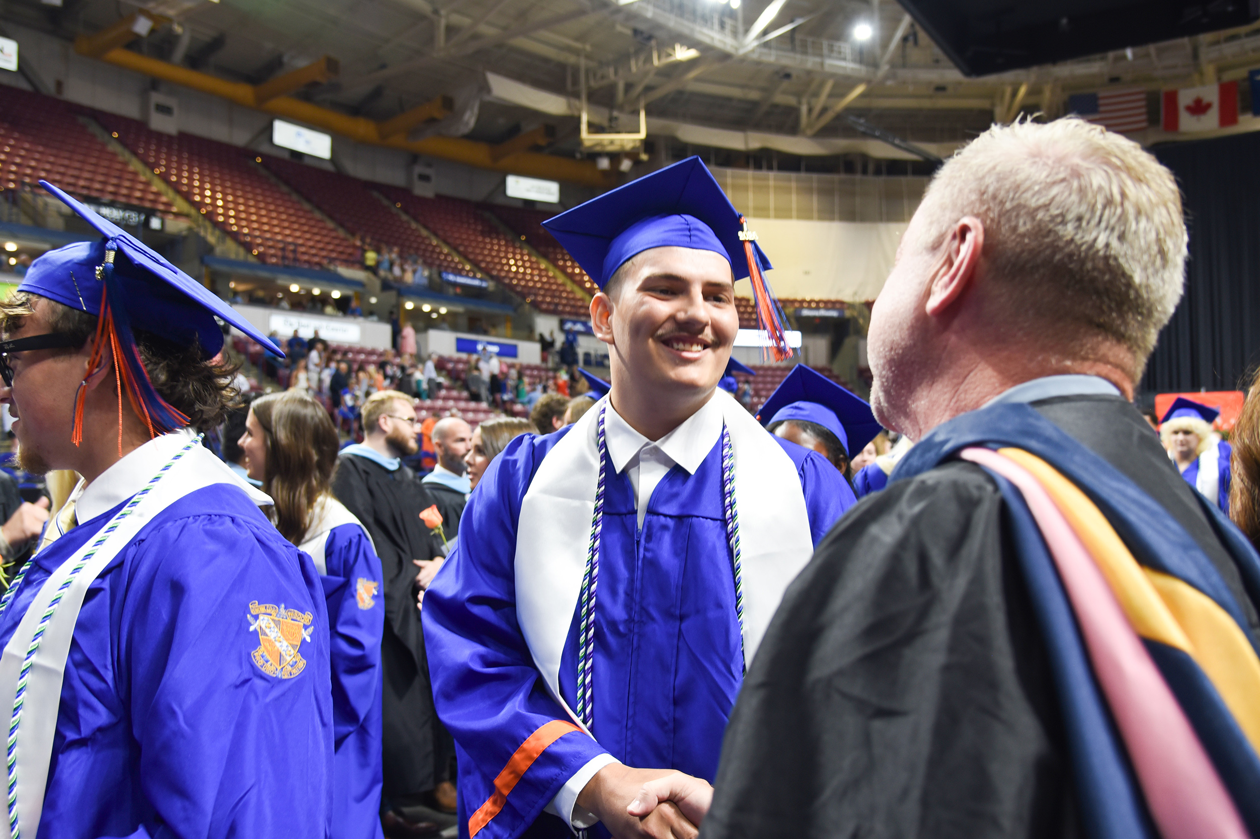 Two students in blue caps and gowns, smiling. An adult in black and pink gown talks with them.