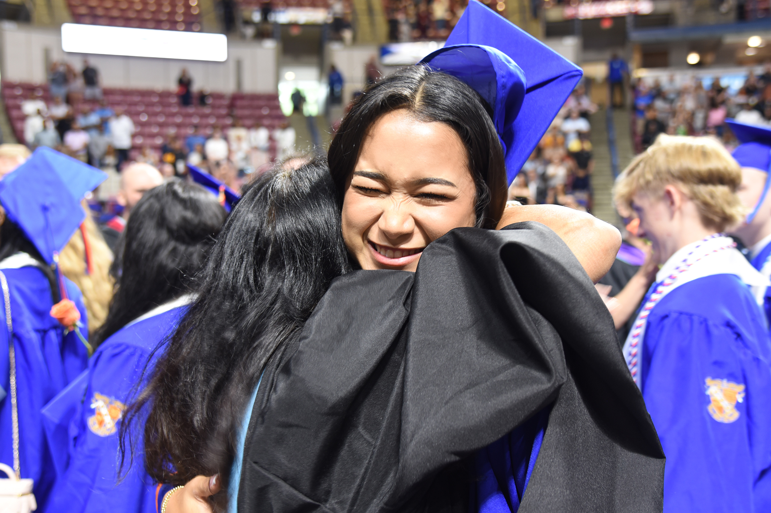 A group of graduates in blue gowns and caps, one smiling woman hugging another, in an auditorium.