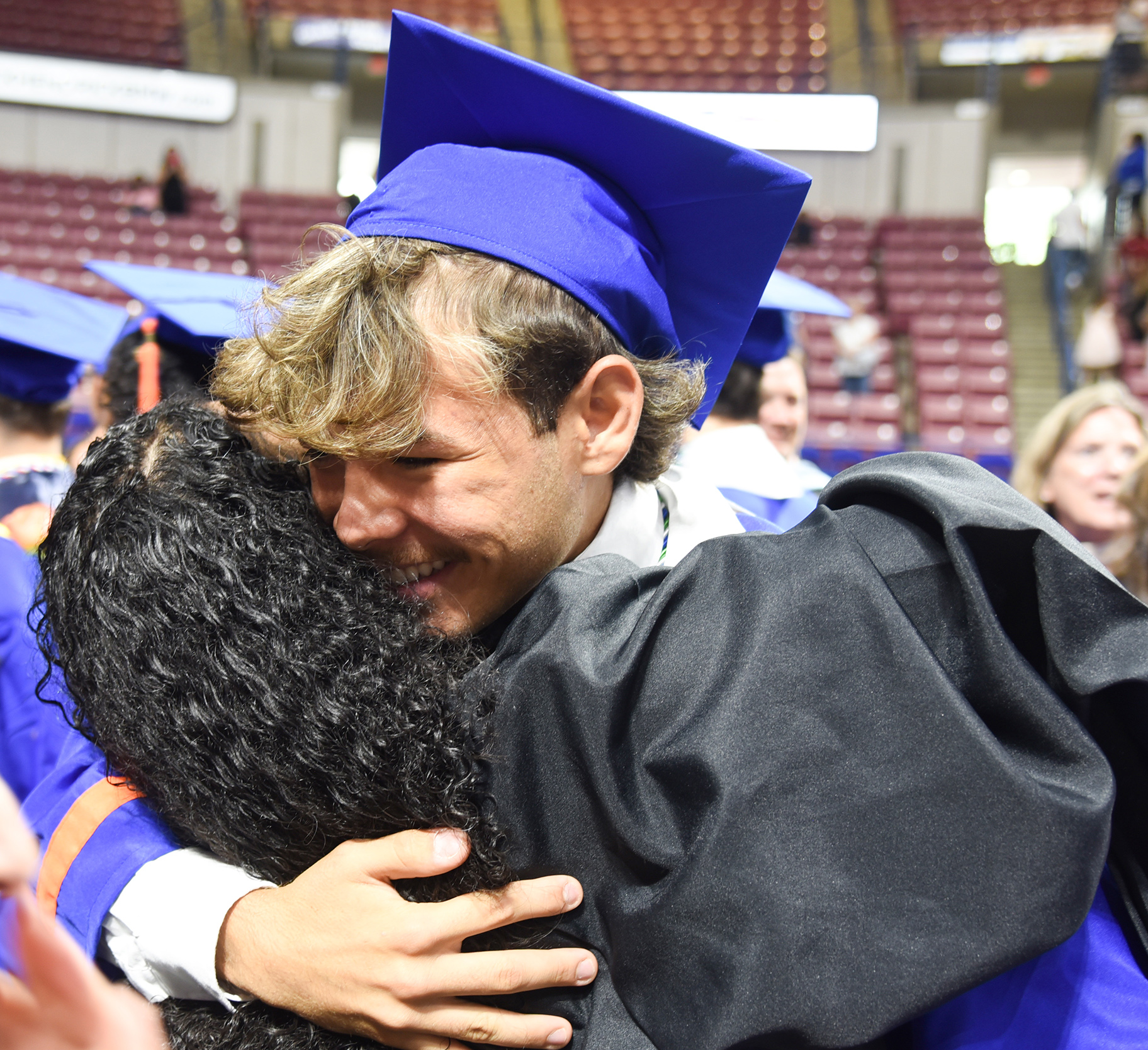 A man in a blue graduation cap and gown embraces another person with curly hair.
