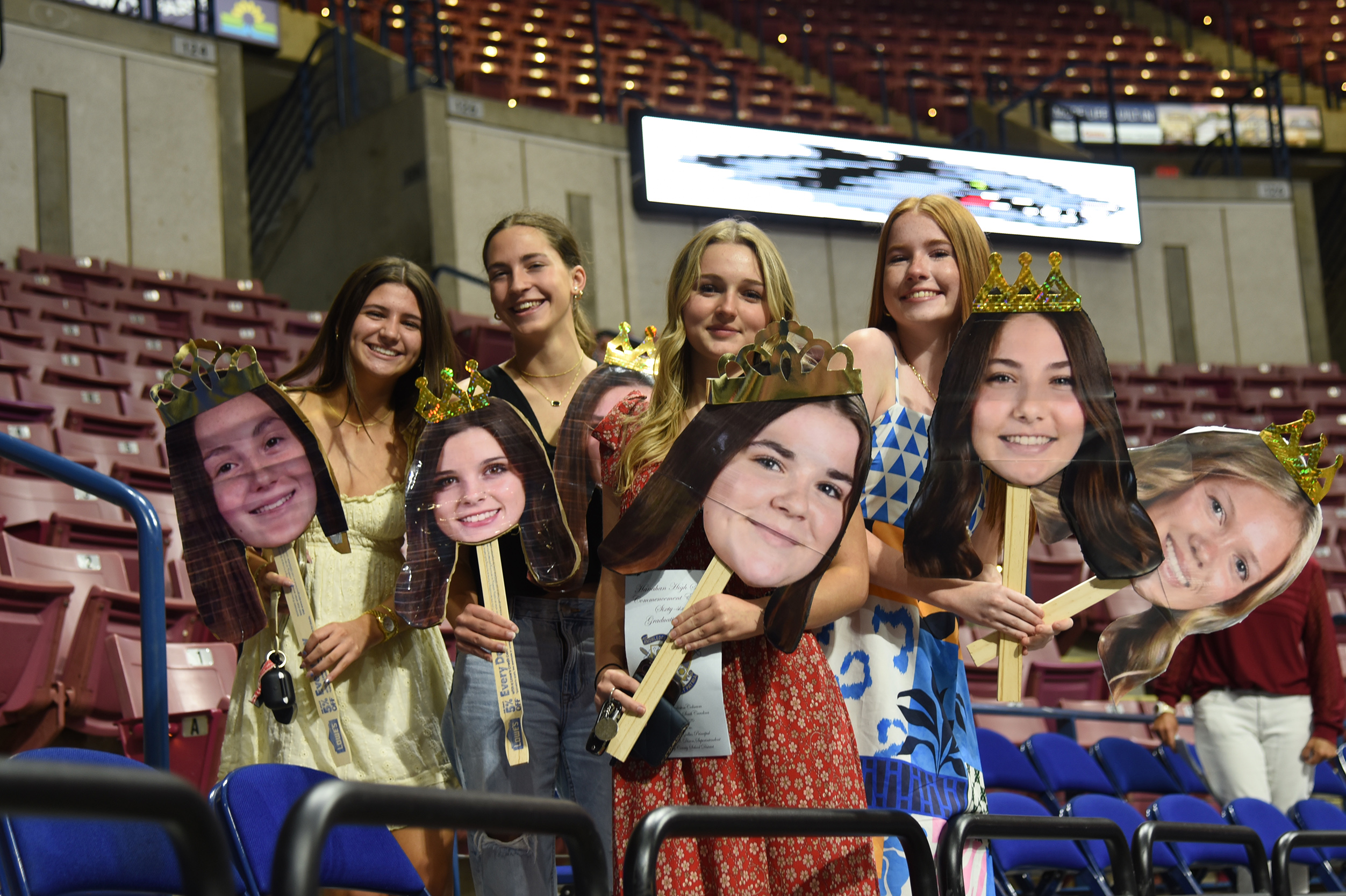 A group of women holding cutouts of faces while standing in an arena. Empty stadium seating and metal railings surround them.