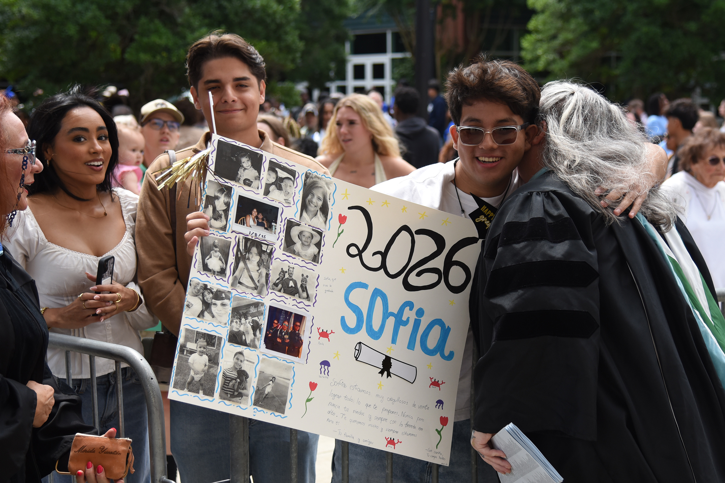 A group of people in a crowd. A man holds a poster with photos and the words "2026 Sofia" in blue.
