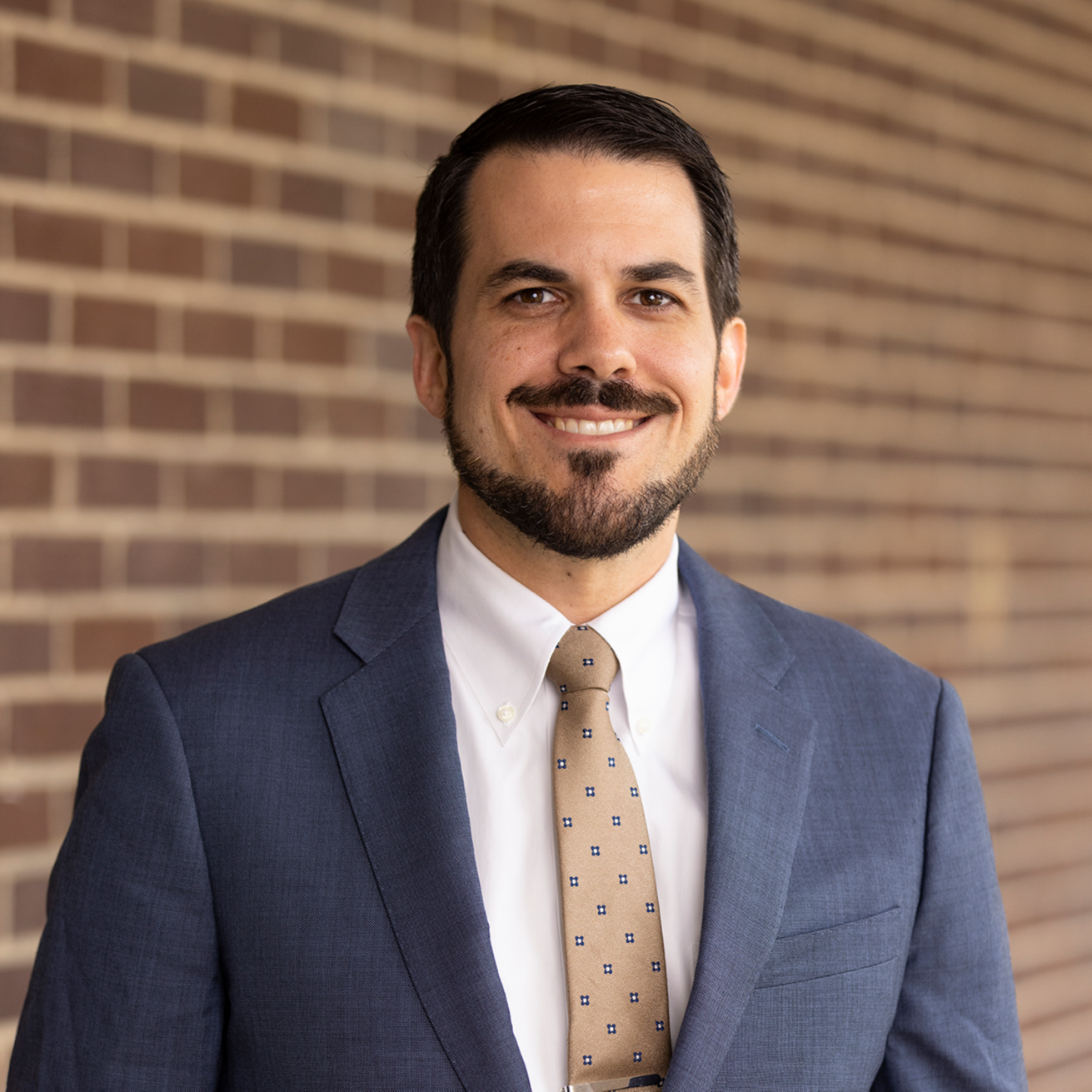A man with a beard and mustache smiles, standing in front of a brick wall. He wears a blue suit with a polka-dot tie.