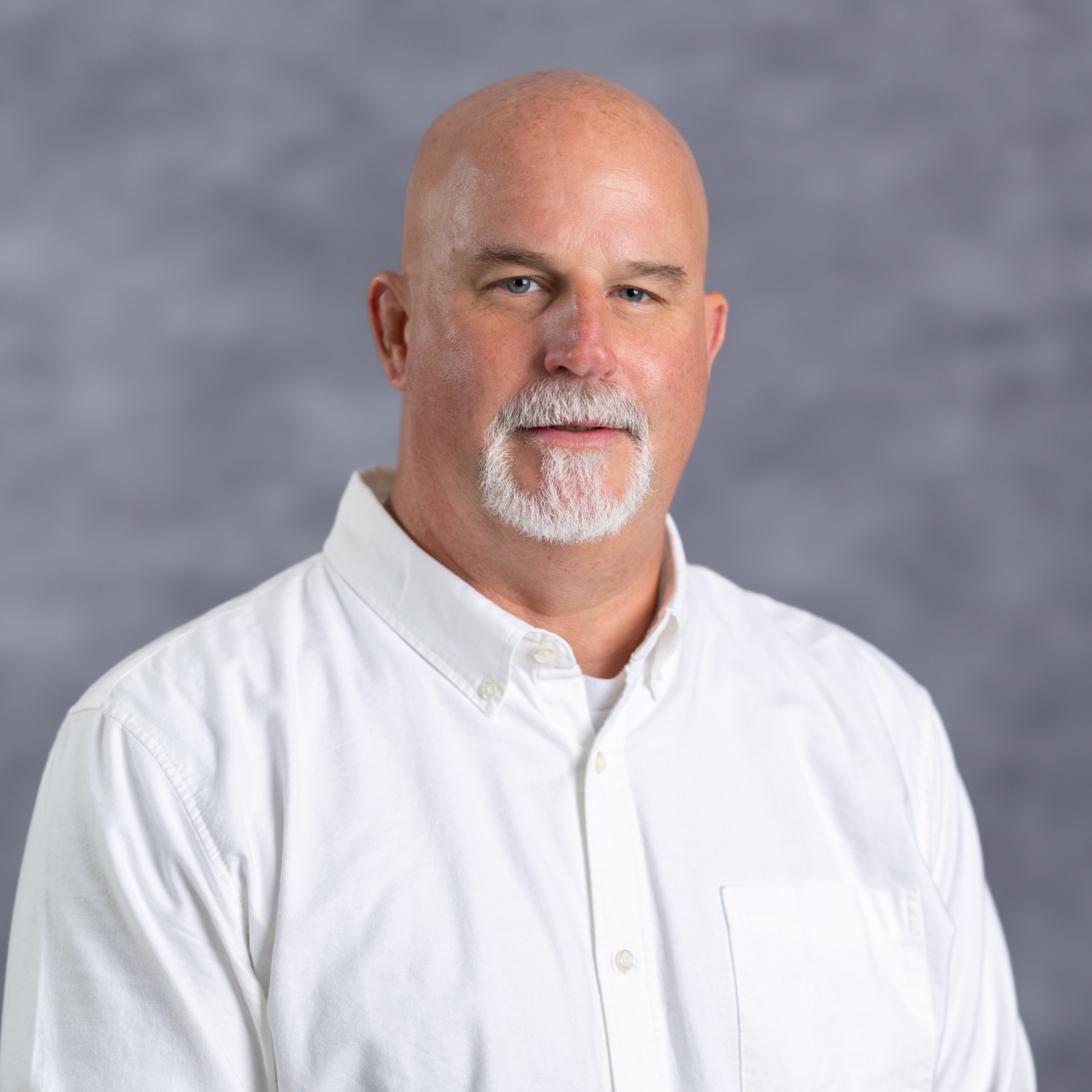 A man with a beard and mustache, bald, wears a white shirt. He stands against a gray background.