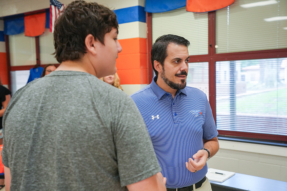 Two men, one in a blue polo shirt, the other in gray, face each other in a room with flag decorations and windows.