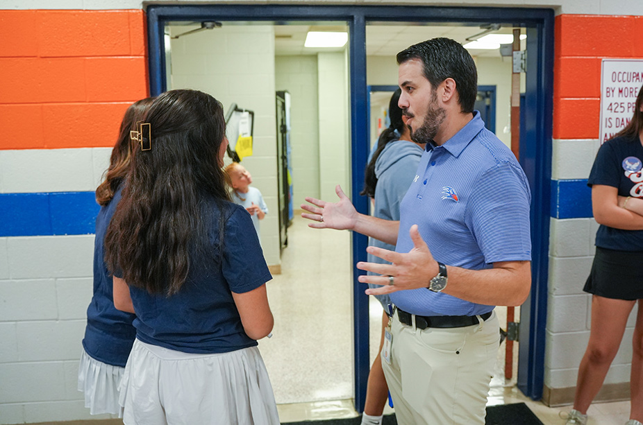 Two women and a man talk in a school hallway with colorful walls, doors, and a carpeted floor.
