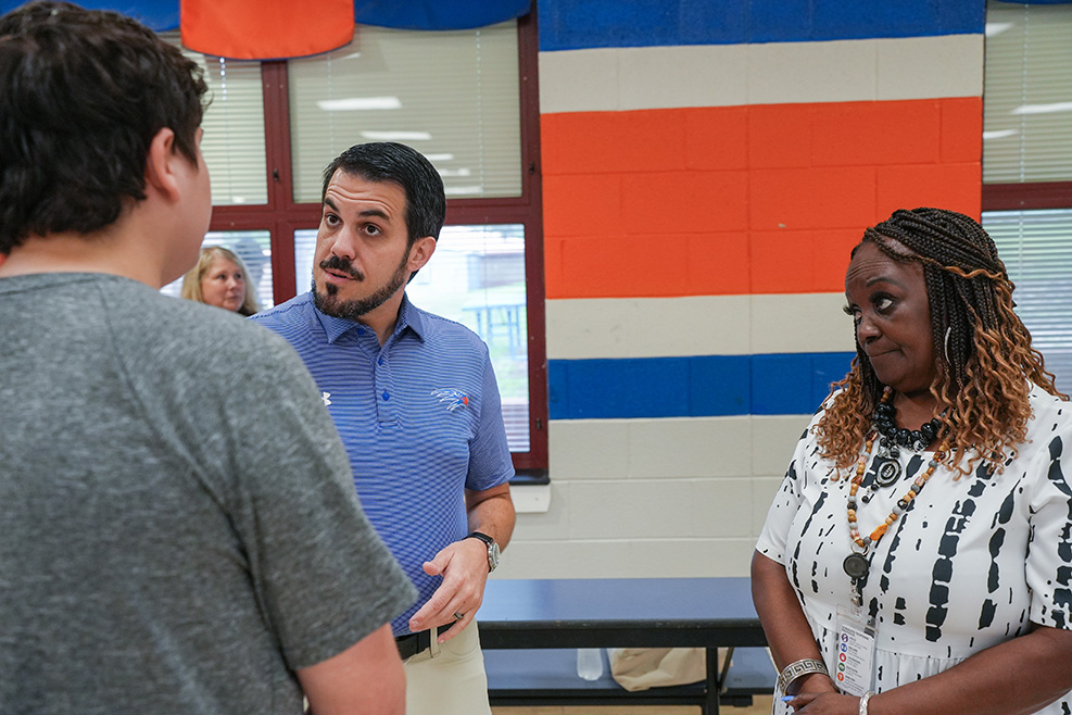Three people in a room. A man in a blue shirt speaks to a woman with curly hair.