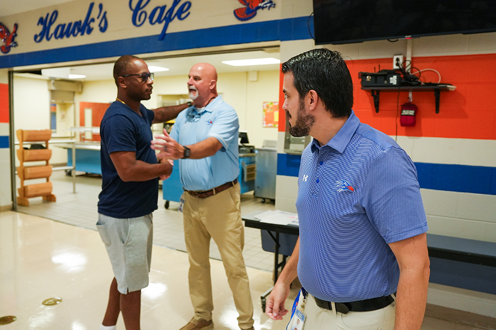 Three men in light blue, blue, and khaki shirts shake hands in a cafeteria with a red, white, and blue wall.