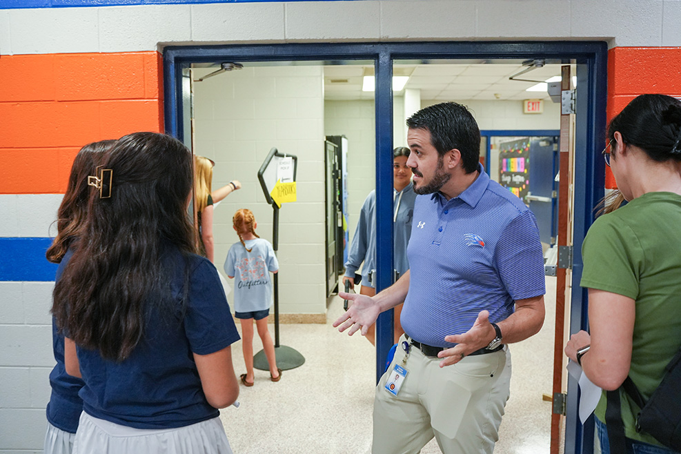Adults and children stand in a hallway. A man speaks to two women. Bright orange and white walls.