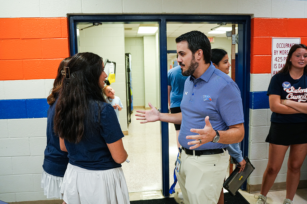 A man in a blue shirt gestures while speaking to a woman in a white skirt. Other people stand nearby.