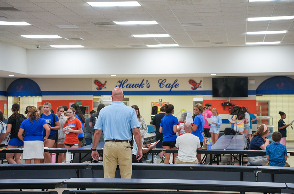 A cafeteria with a banner reading "Hawk's Cafe" and lights on the ceiling. Many people are gathered.