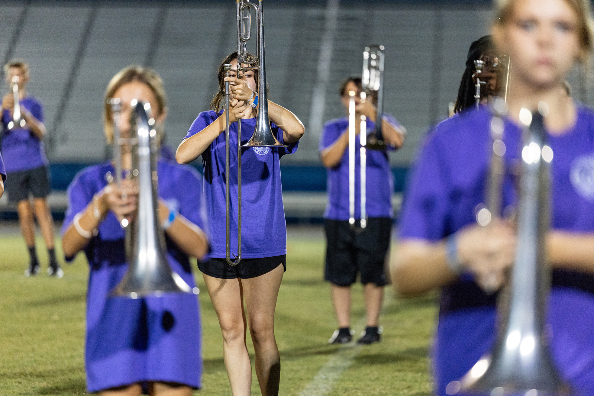 A group of individuals in purple shirts and black shorts playing trombones on a grassy field.