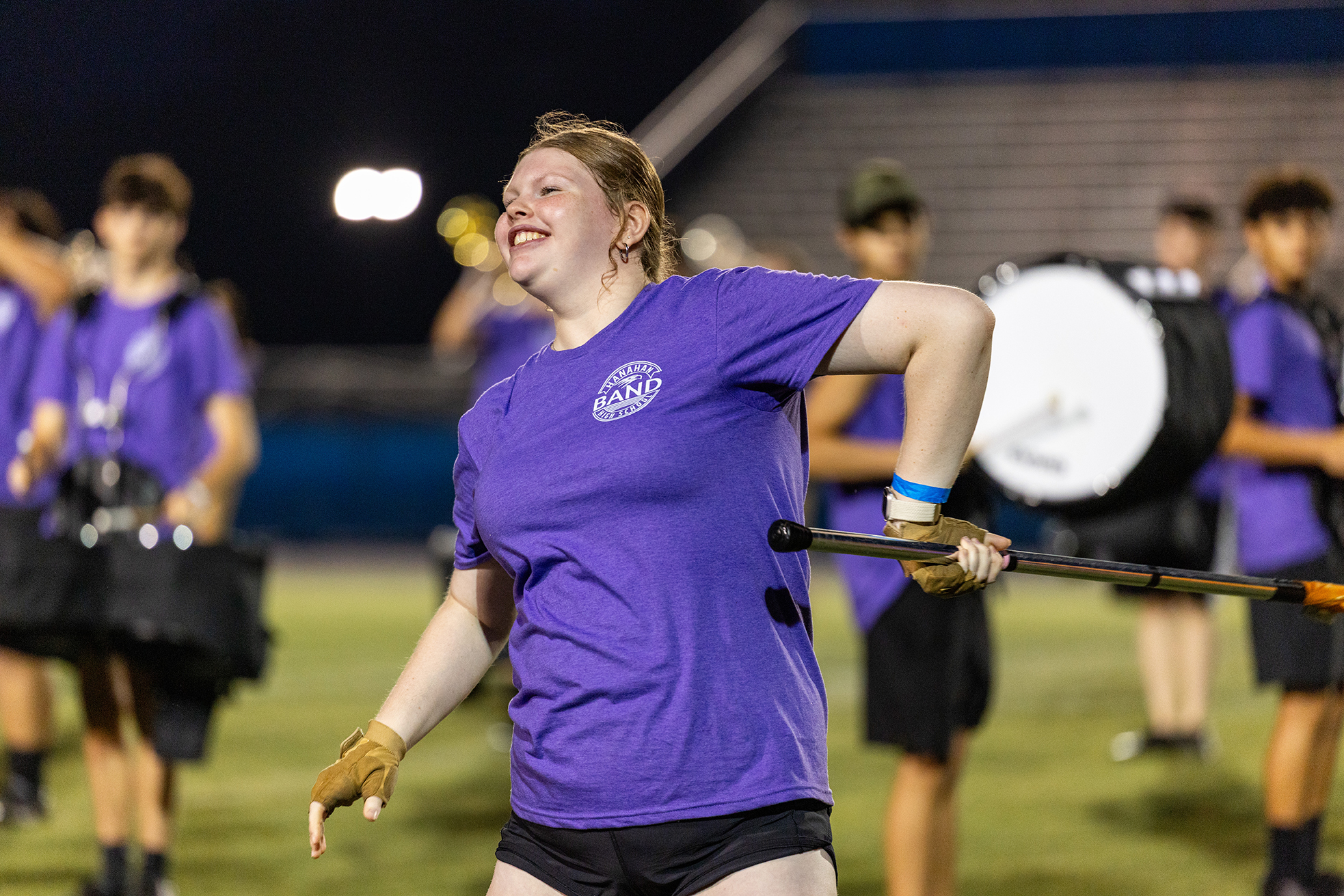 A woman in a purple t-shirt and black shorts twirls a baton while smiling. Several others stand behind her on a field.