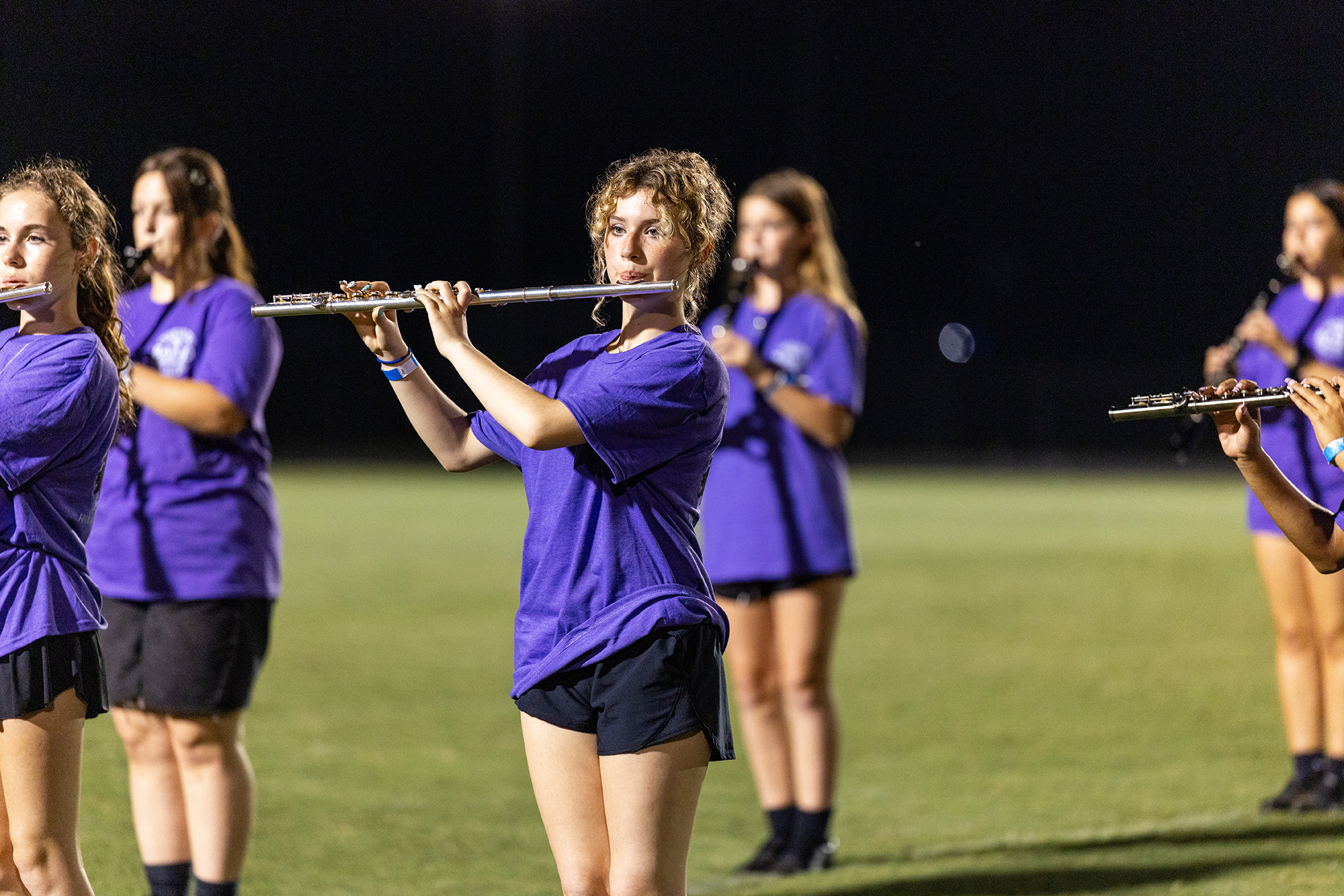 A group of women in purple shirts and black shorts play flutes on a grassy field.
