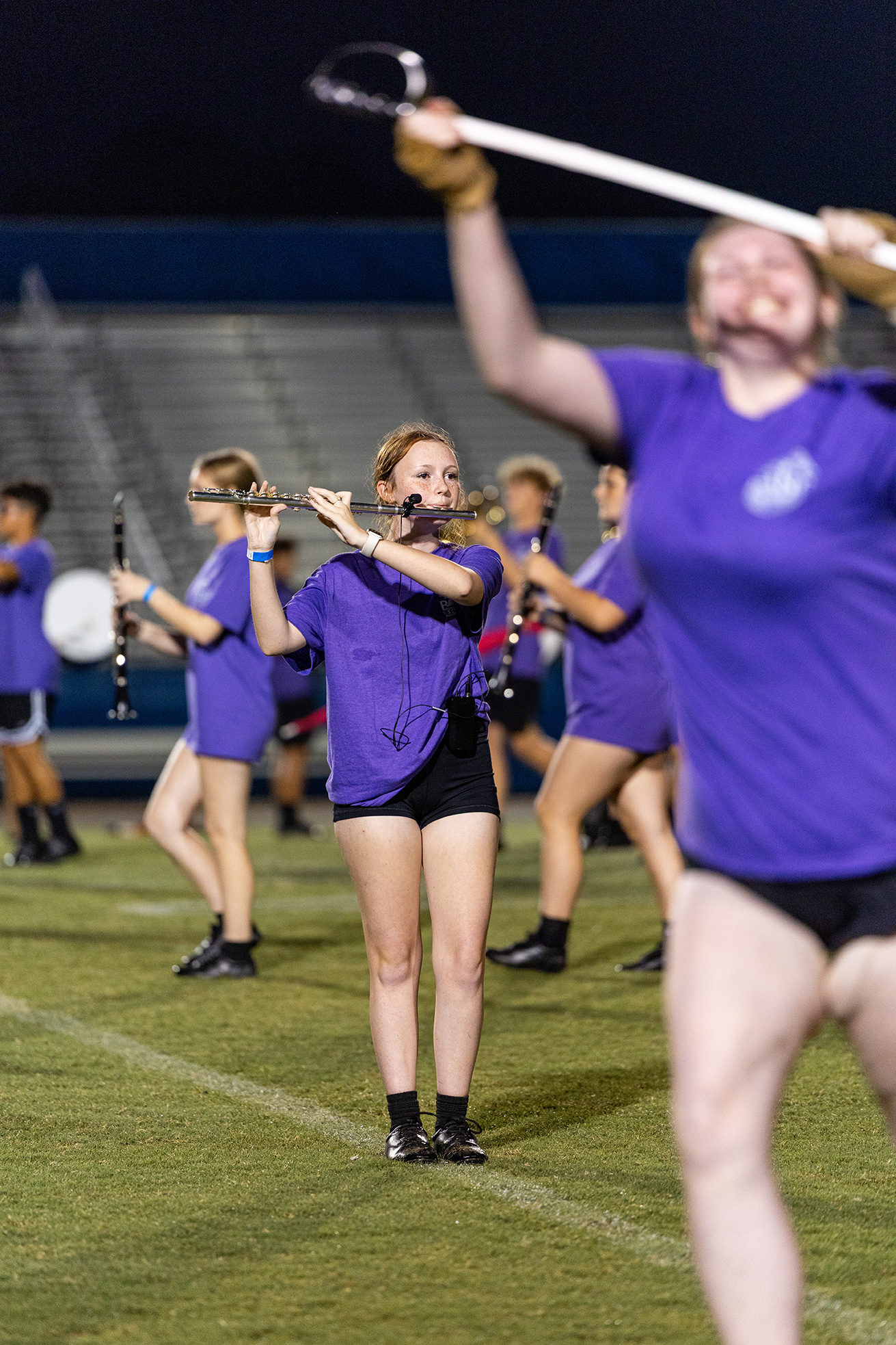 A band with purple tops and black shorts performs on a field. A woman holds a flute, while others hold a drumstick and a baton.