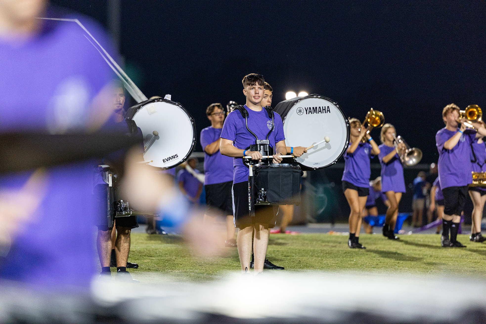 Musicians in purple uniforms stand on a field with instruments, including trumpets, drums, and trombones.