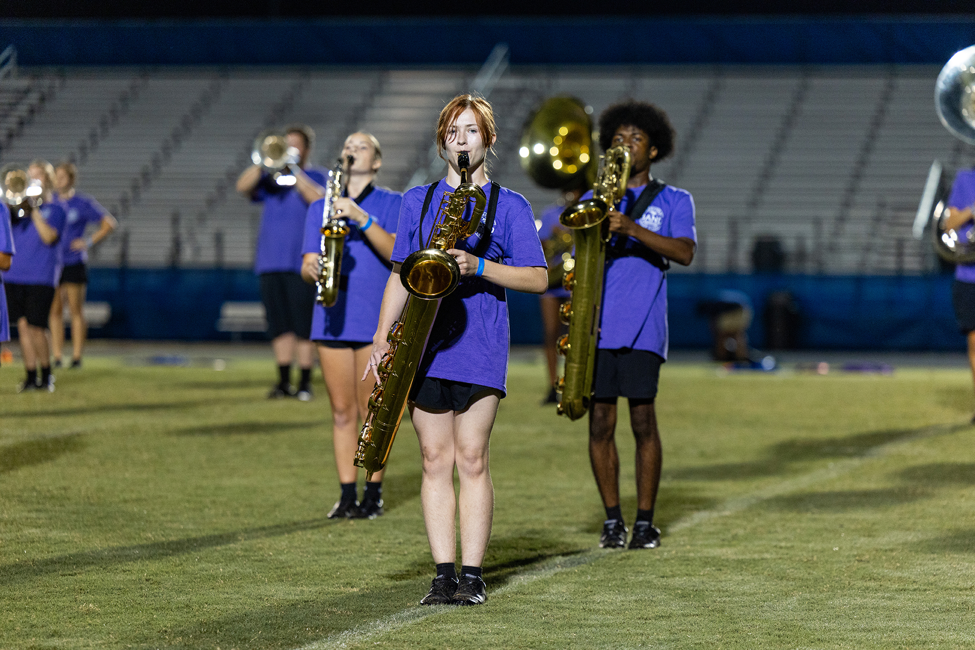 A group of musicians in purple shirts and black shorts perform on a field with large brass instruments.