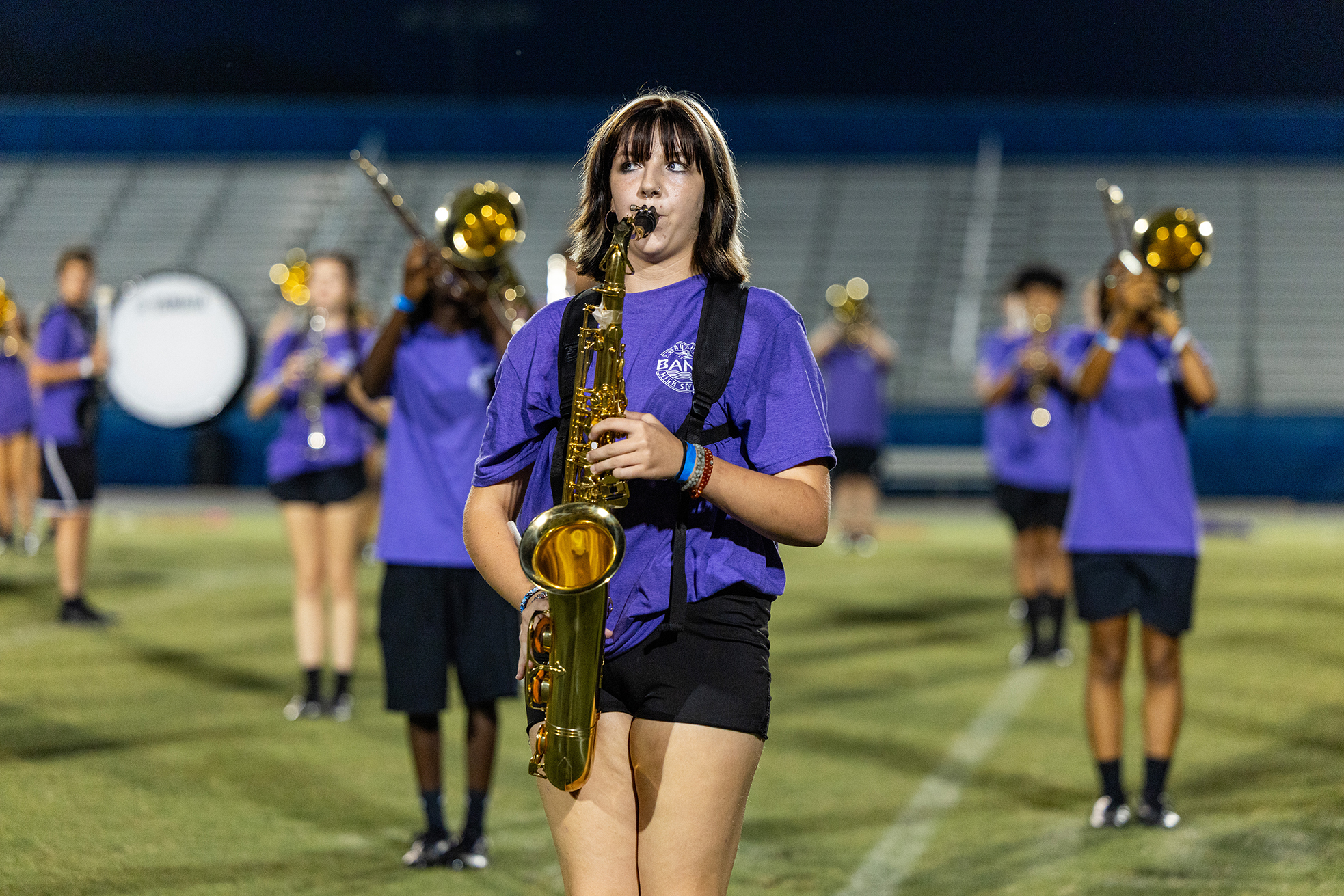 A group of students in purple shirts and black shorts perform on a field at night, with one holding a saxophone.