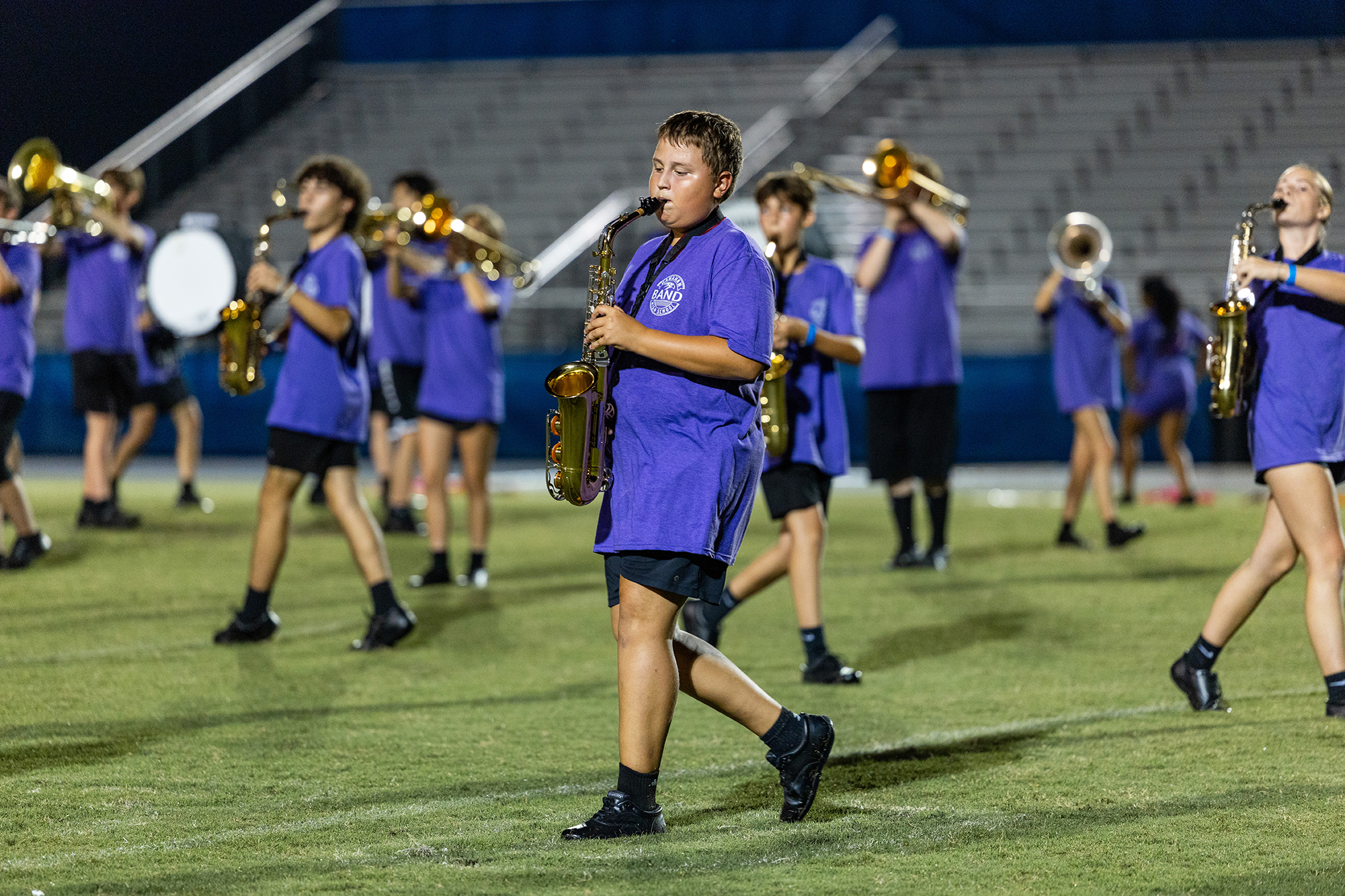 A school band practices on a football field at night, with students in blue shirts playing instruments, including saxophones and trumpets.