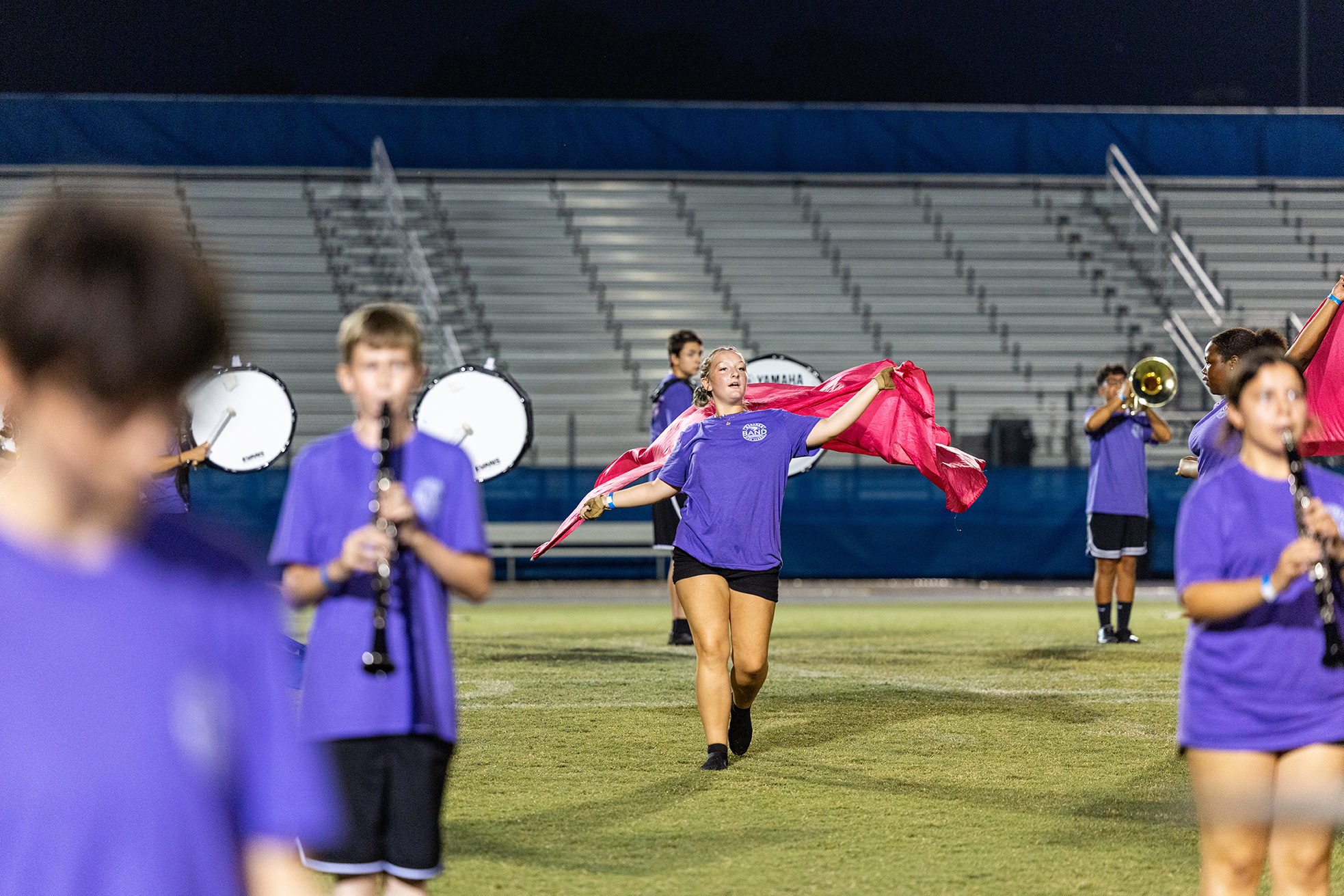Band members in purple tops and shorts on a field, with one holding a pink flag. Empty bleachers in the background.