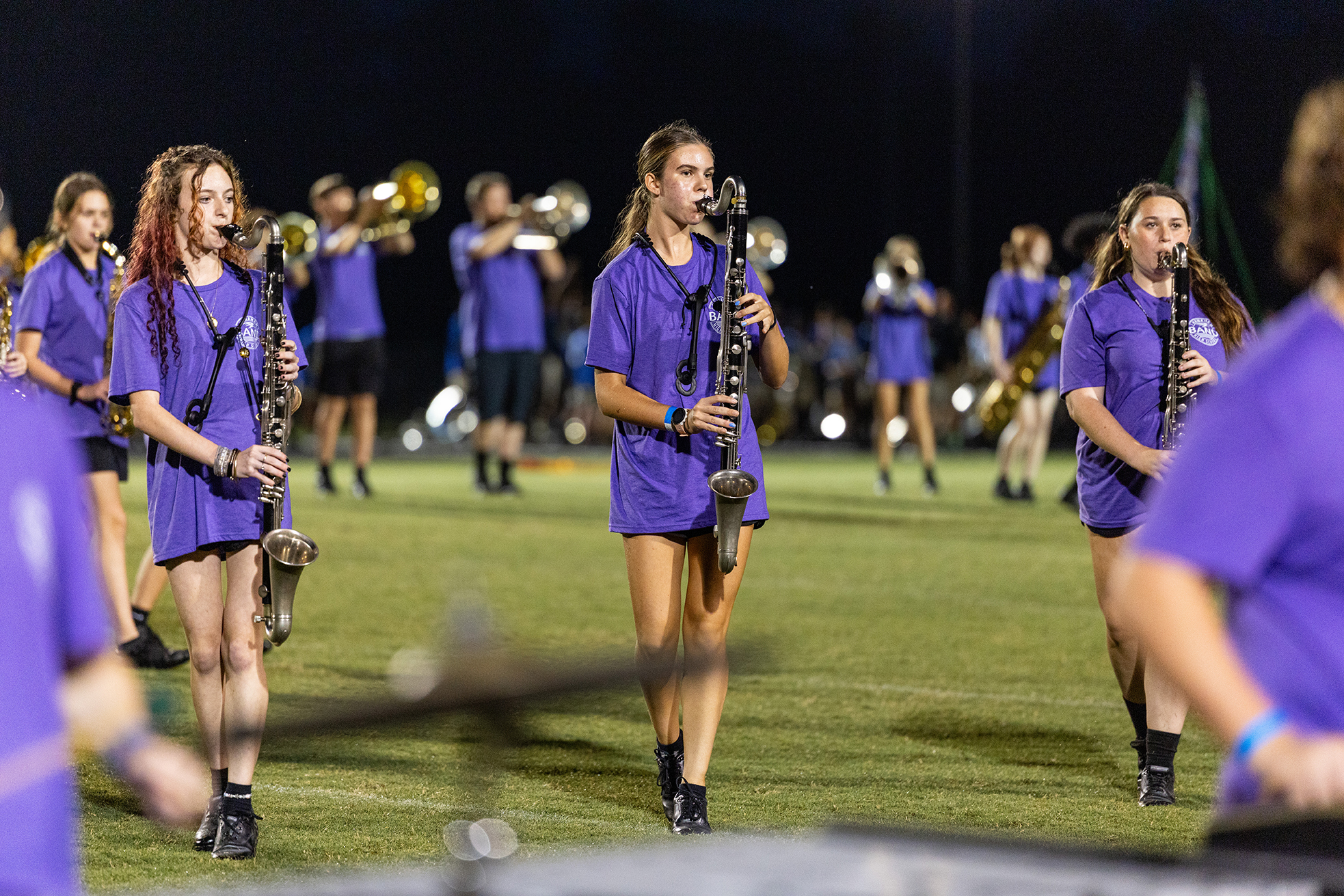 Musicians in purple uniforms, some playing instruments, walking on a field with a blurred audience in the background.