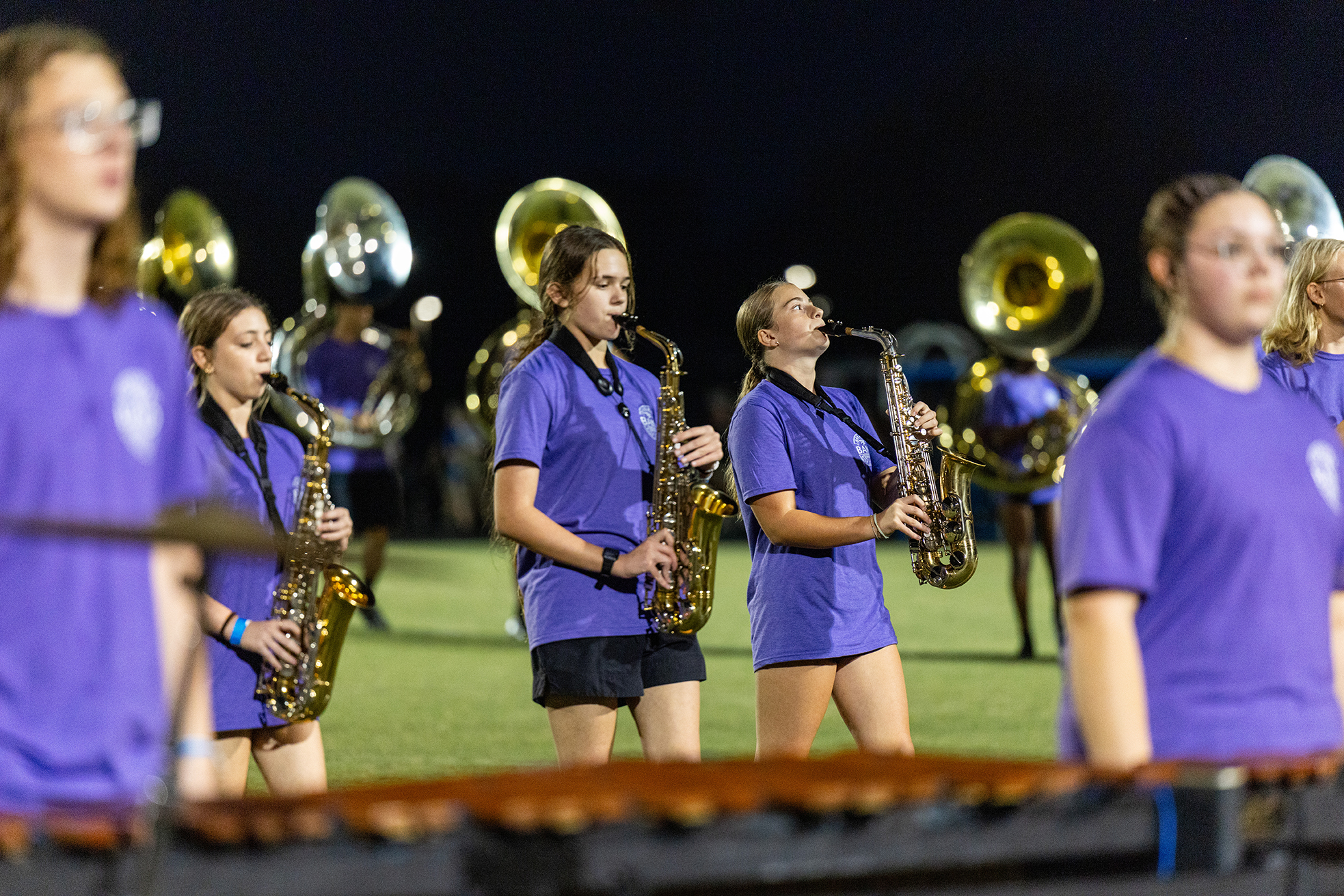 A group of women in purple uniforms playing saxophones on a field at night, with a xylophone in the foreground.