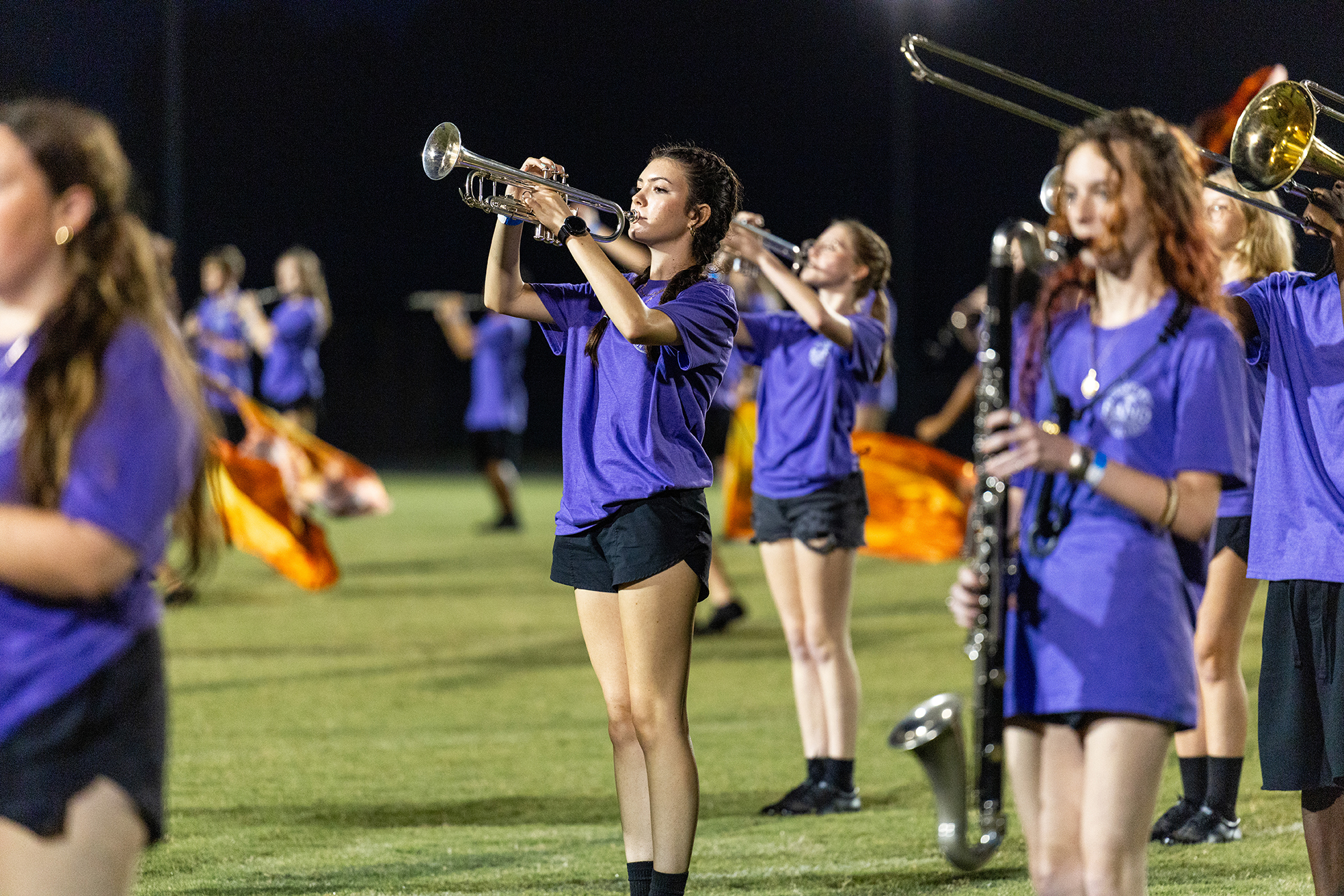 A group of young women wearing purple shirts and black shorts perform with instruments on a field.
