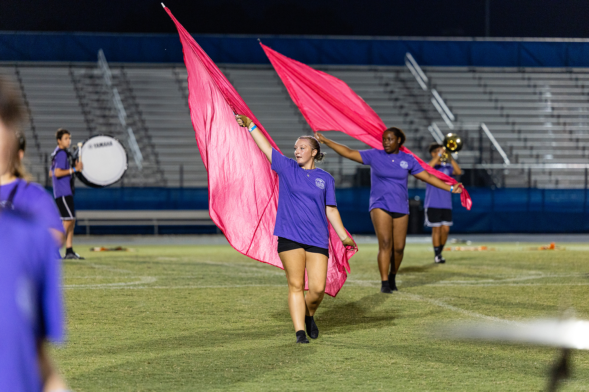 Several people in purple shirts and black shorts on a field at night. One person holds pink flags.