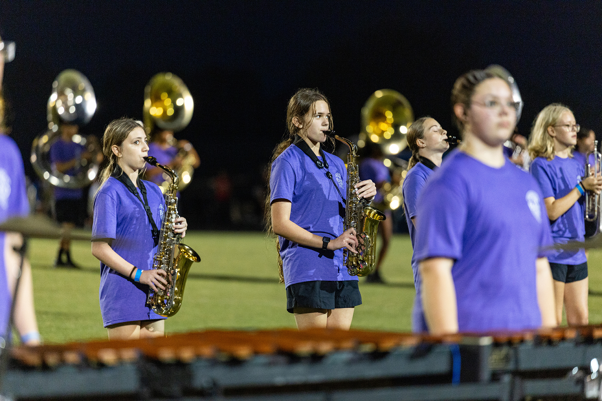 A group of musicians in purple shirts, playing saxophones and a marimba, stands on a field.