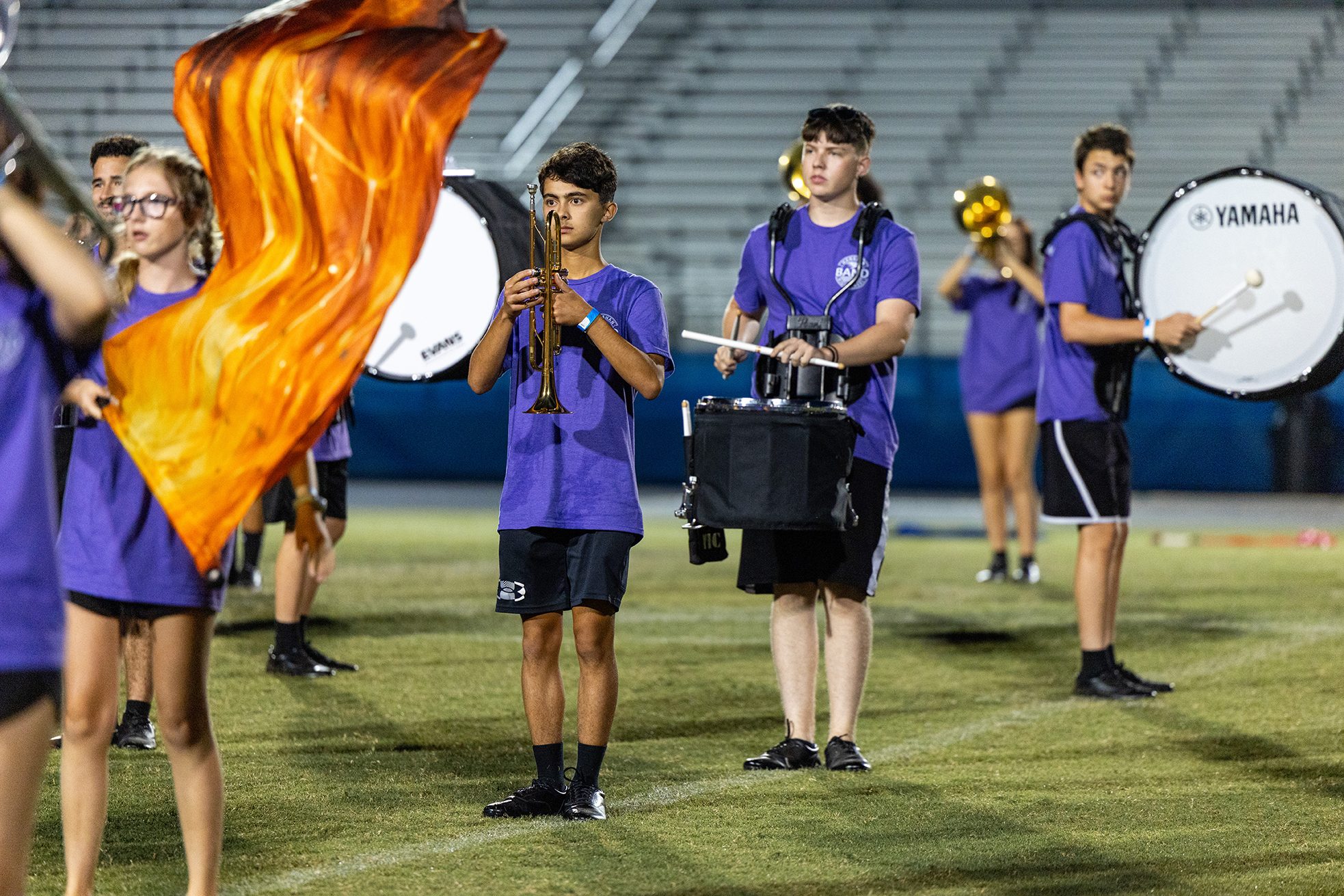 Group of band members on a field wearing purple shirts. Some hold drums and others have instruments in their hands.