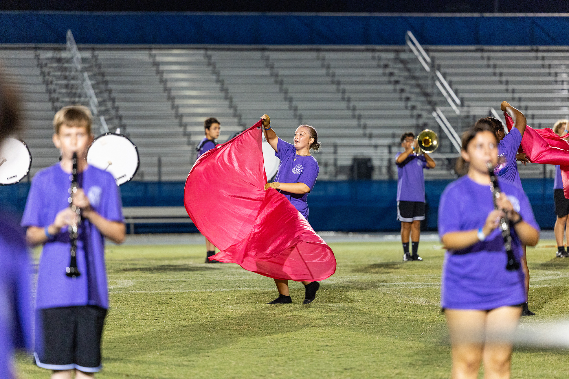 A group of children perform on a field with instruments. A girl in front holds a pink fabric.