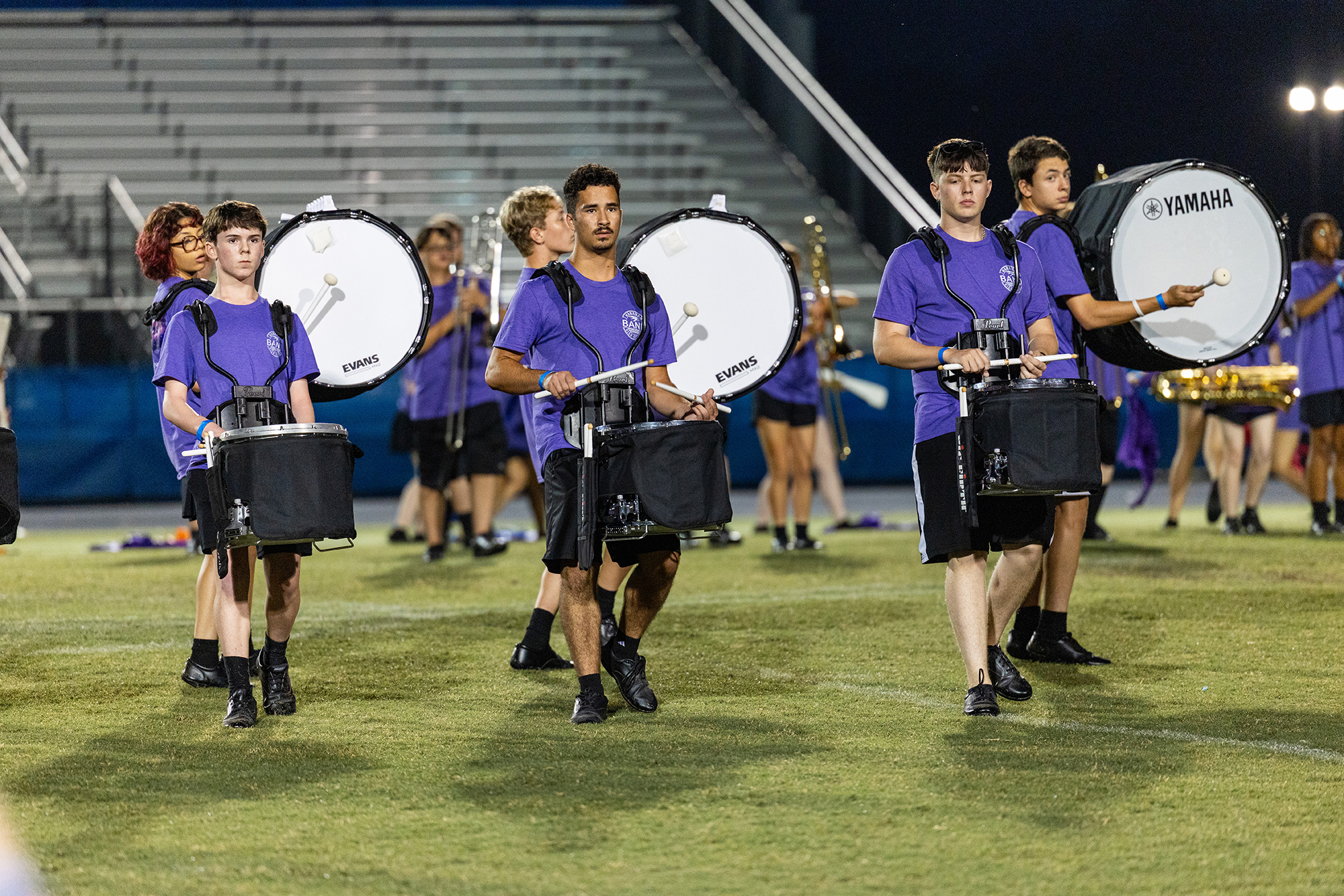 A group of young people in matching uniforms playing drums on a grassy field at night.