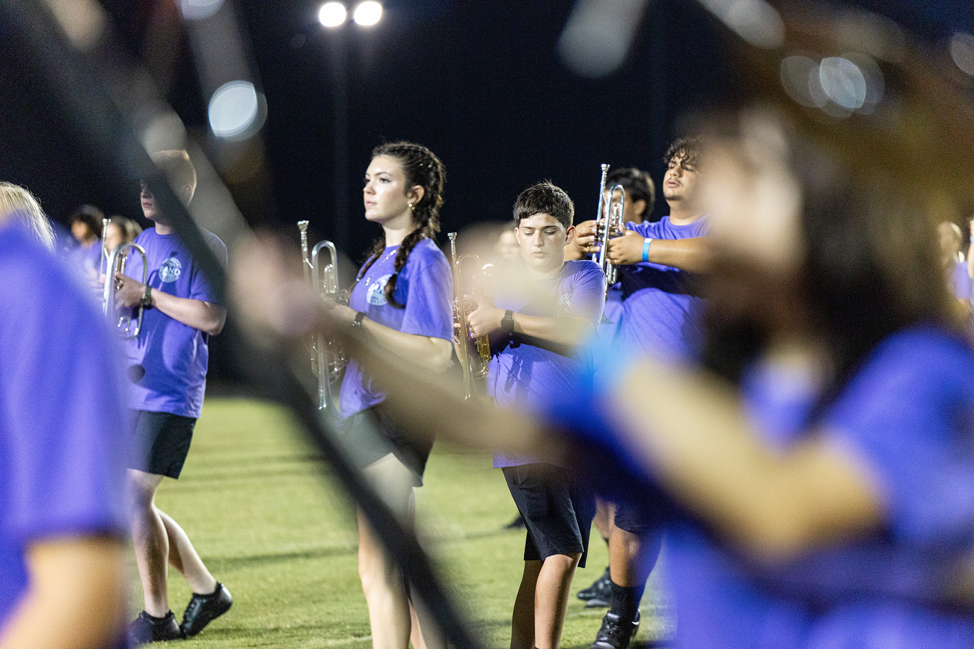 A group of people in blue shirts and shorts on a field, playing instruments. They are illuminated by lights.